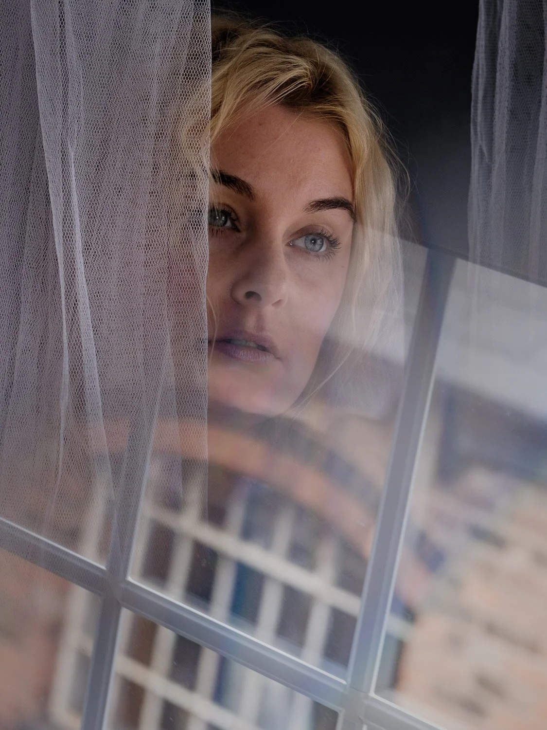 A woman with blonde hair and blue eyes looks through a window with a sheer curtain partially covering her face.