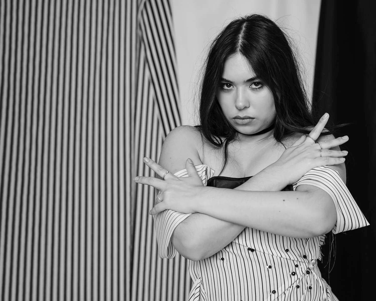 A young woman with dark hair and a serious expression, wearing a striped off-shoulder shirt, crosses her arms in front of her chest in a pose against a background with vertical lines.