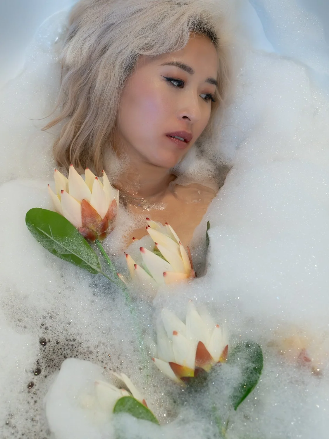 Woman with blonde hair relaxing in a bubble bath surrounded by large flowers and green leaves.