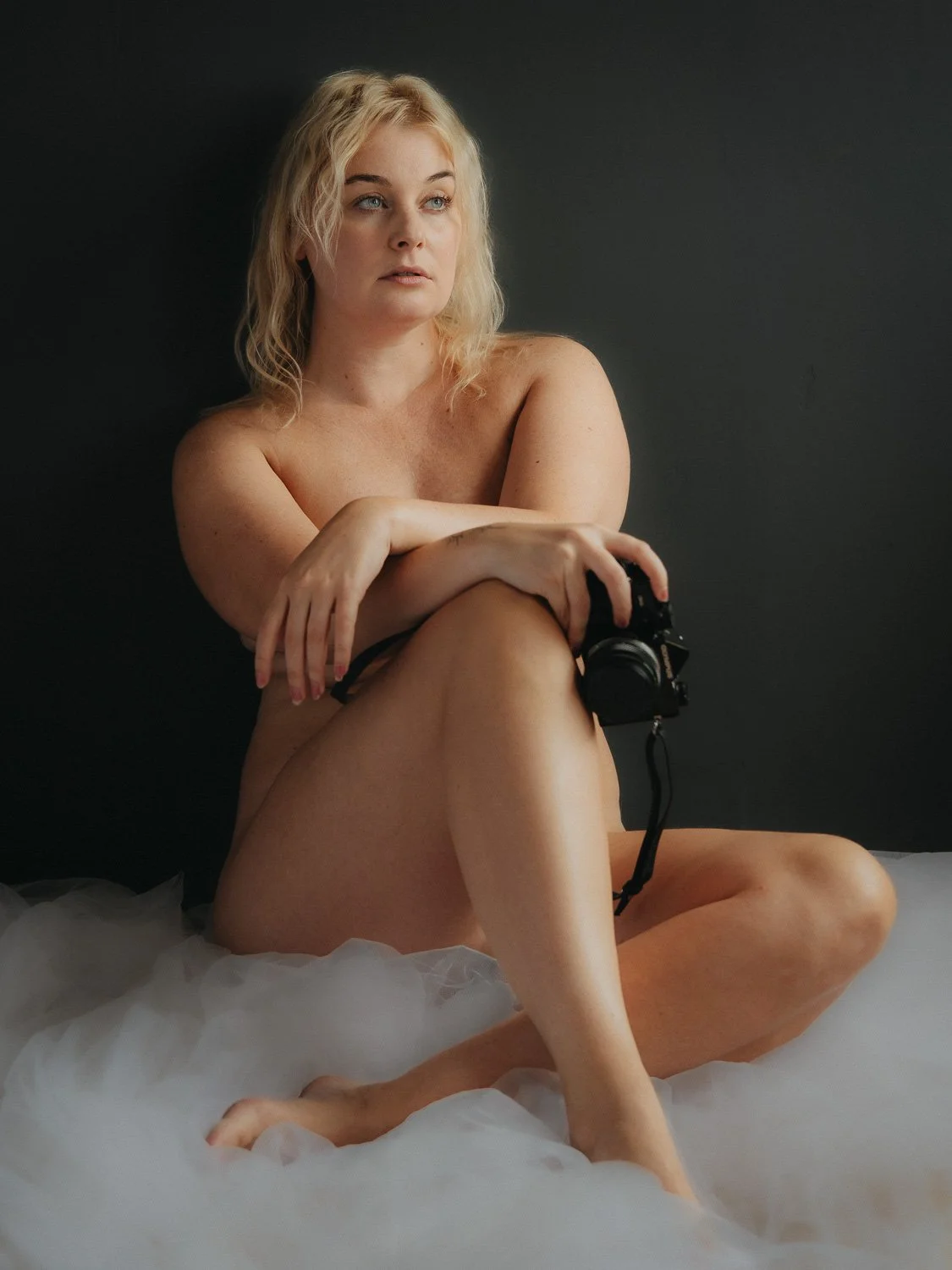 A nude woman with blonde hair sitting on a bed with white sheets, holding a camera, against a dark background.