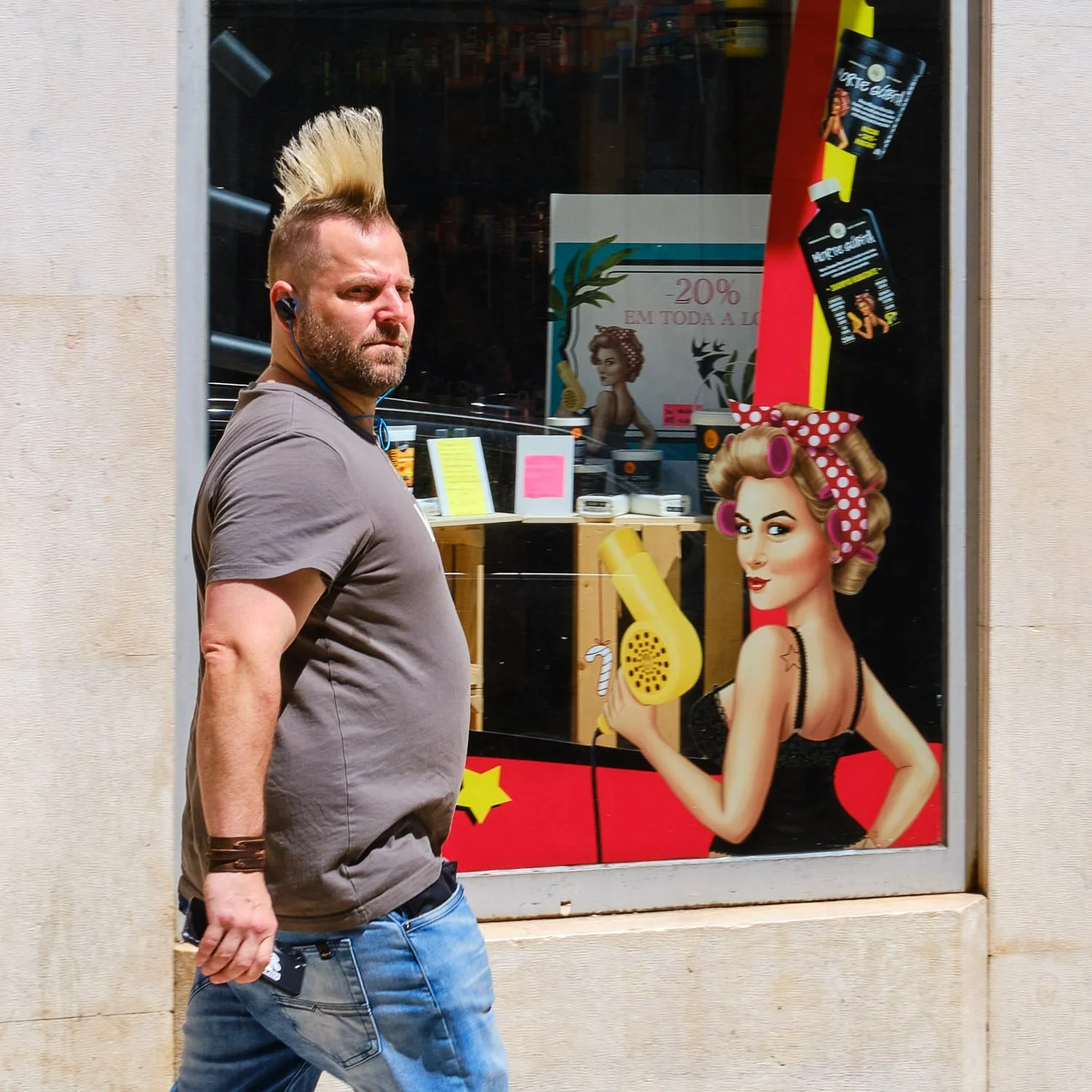 A man with a mohawk hairstyle, wearing a gray t-shirt and jeans, walking past a store window display featuring a vintage-style woman with curled hair and roller curlers, holding a hairdryer.