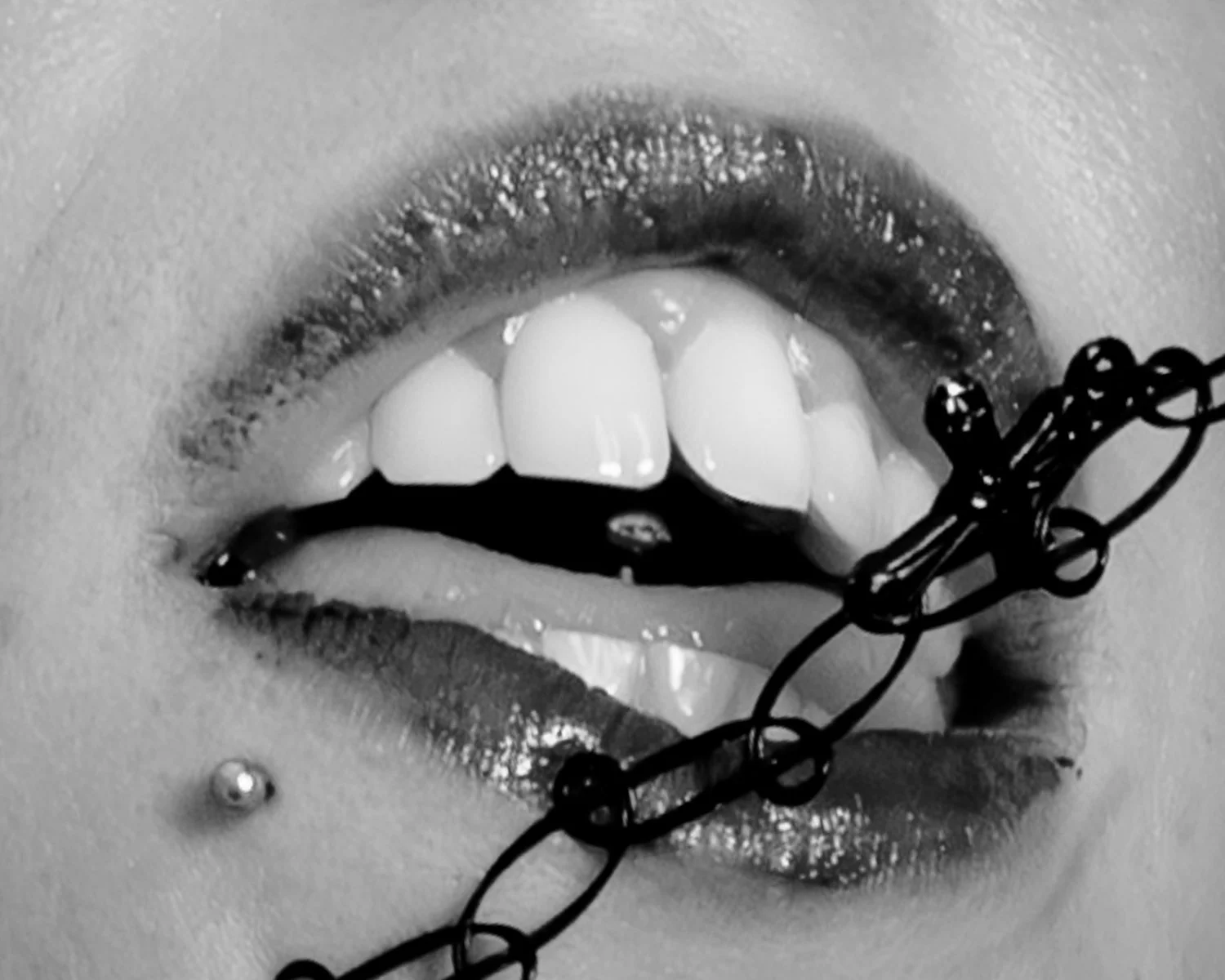 Close-up black and white photo of a person's open mouth with lips slightly parted, showing teeth and tongue, with chain jewelry around the lips and a lip piercing.