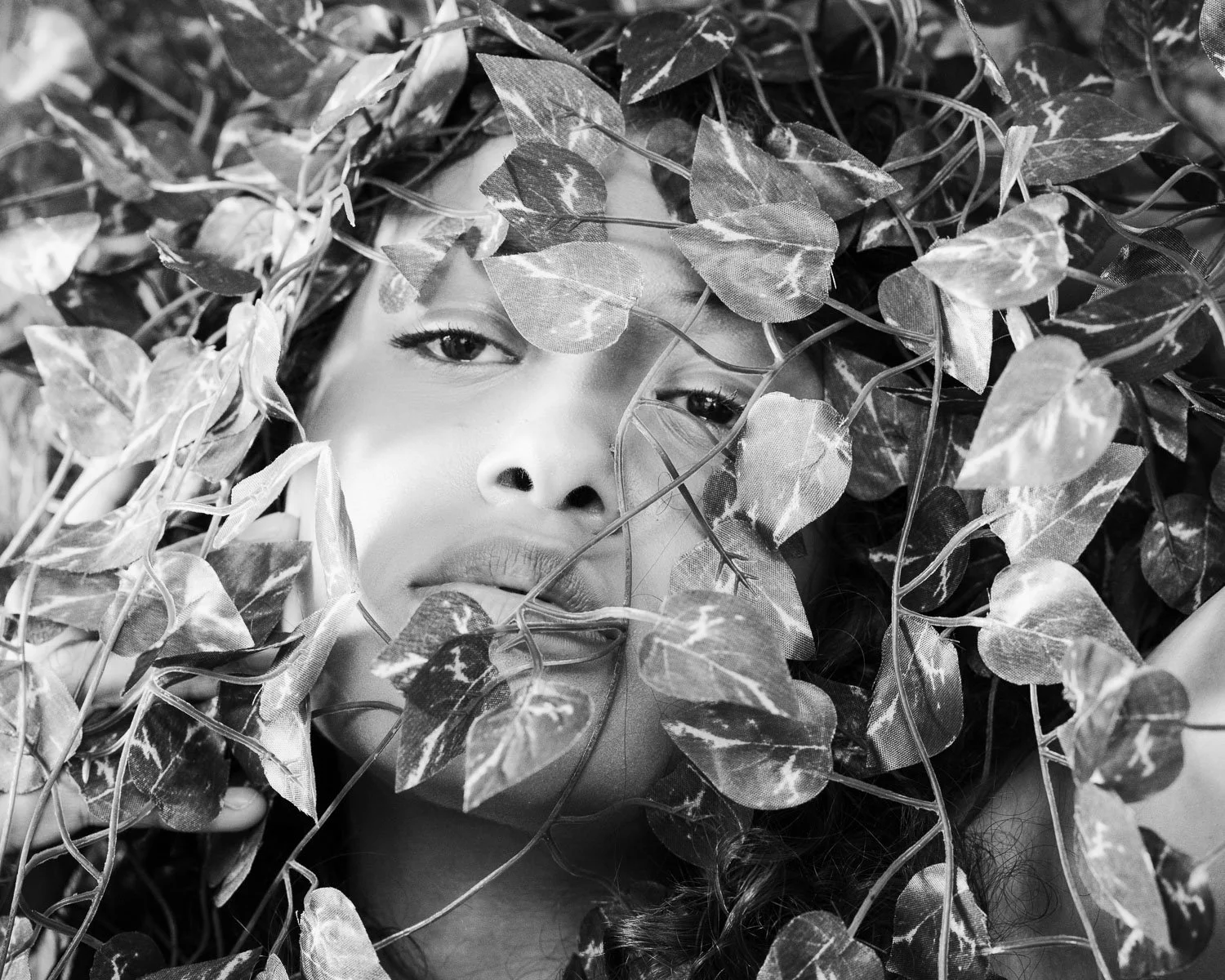 Black and white photo of a woman's face surrounded by and partially obscured by jungle leaves.