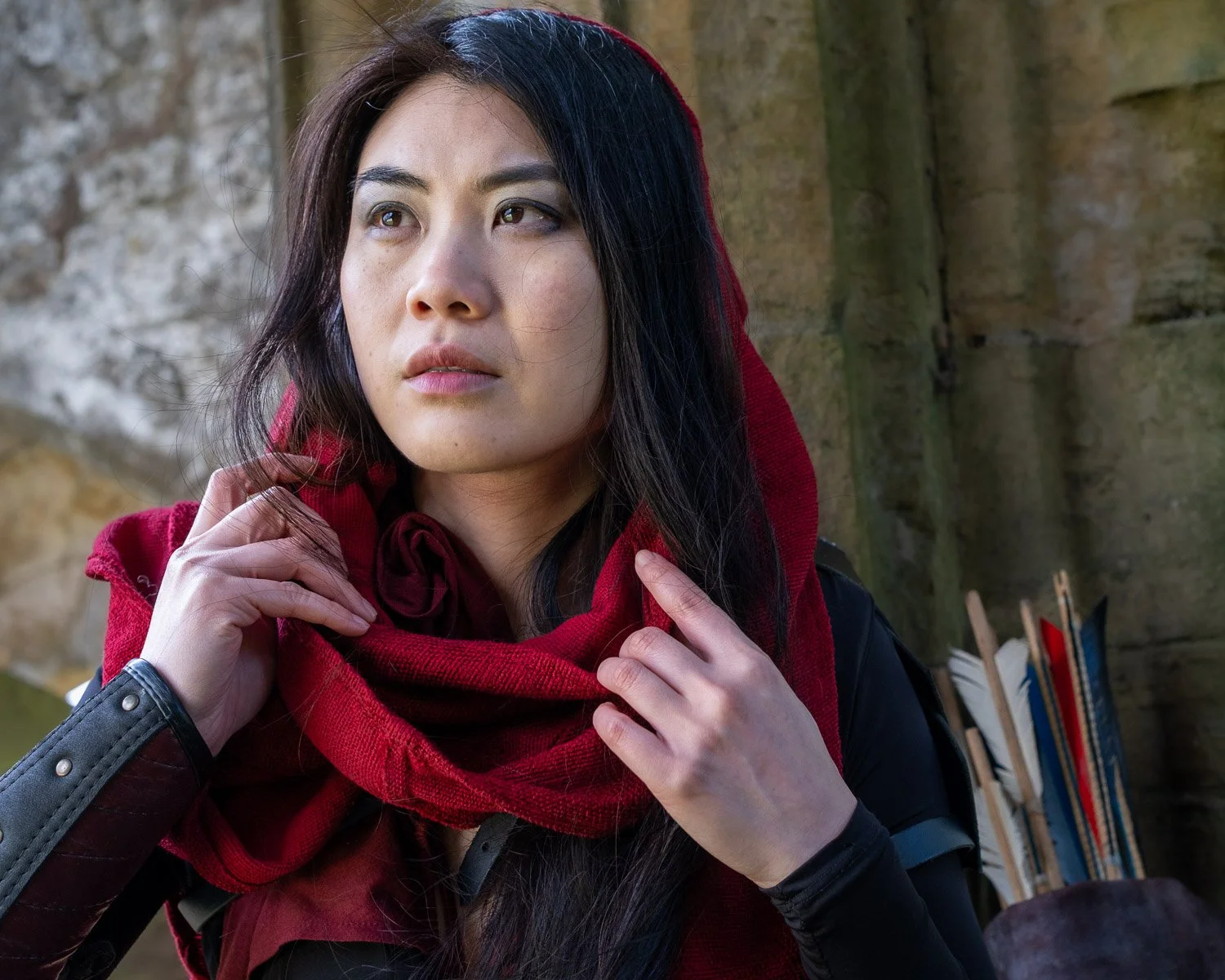 A woman with long black hair and a red hoodie, looking into the distance with a thoughtful expression, sitting outdoors against a stone wall with books in the background.