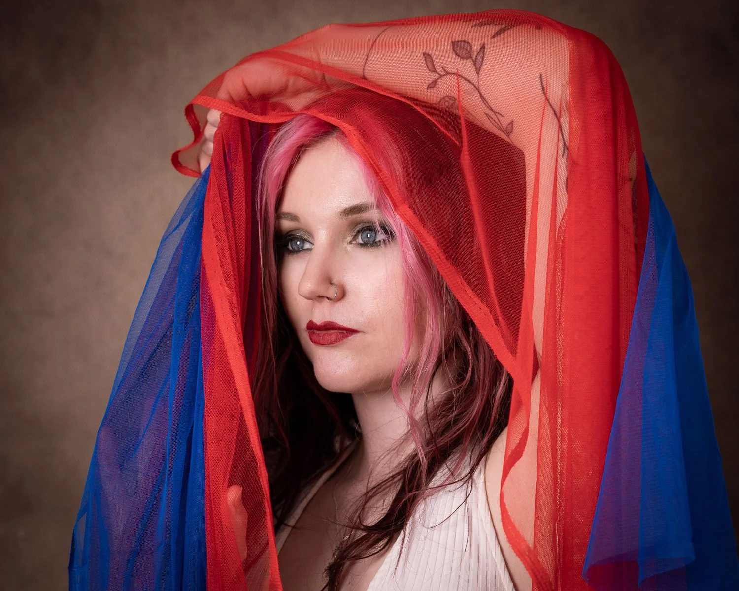 A woman with pink hair and blue eyeshadow is gazing to the side, wrapped in red and blue sheer fabric.
