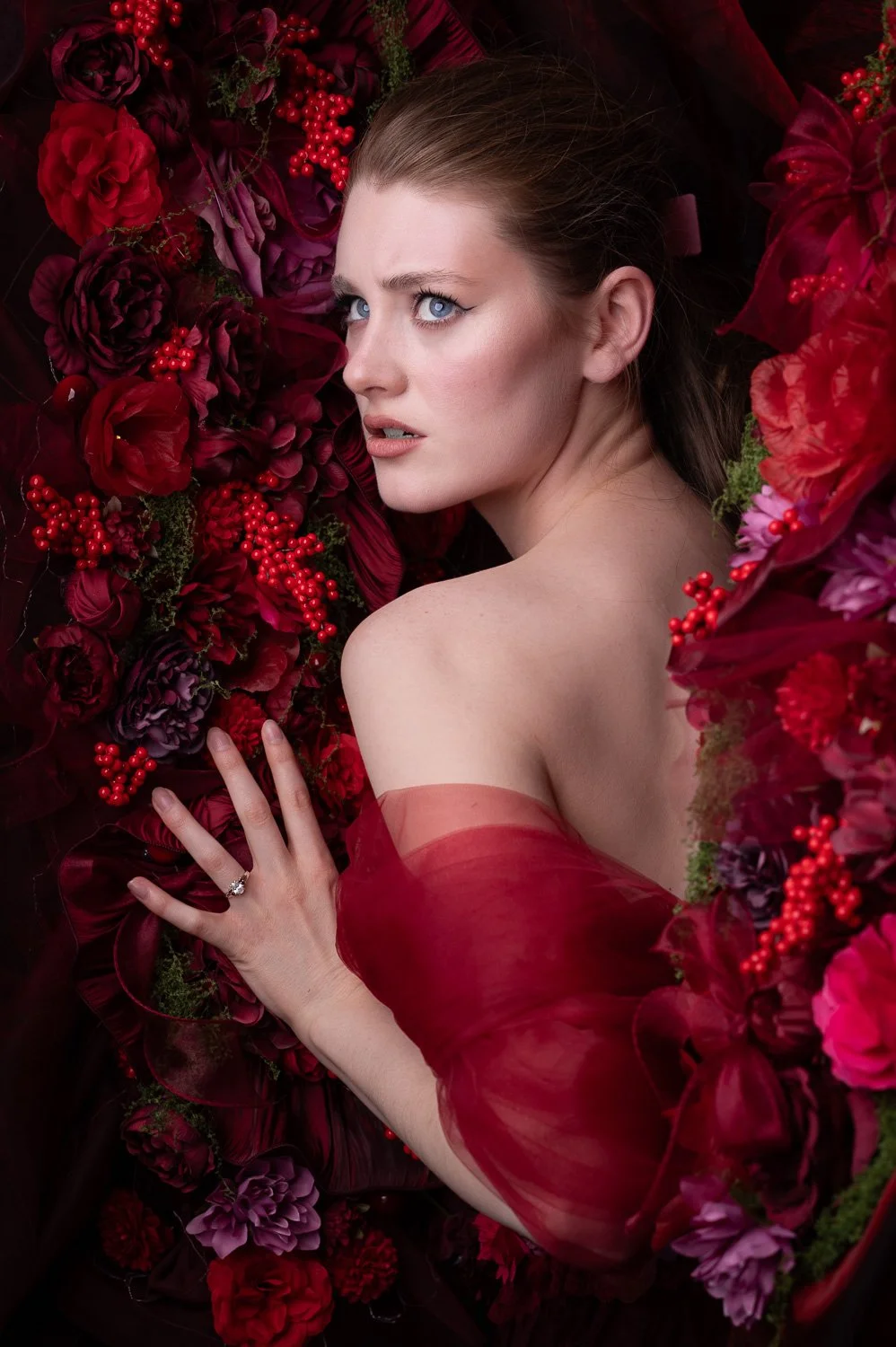 A woman with blue eyes and fair skin lying among deep red and purple flowers, wearing a sheer red off-shoulder dress and a ring on her finger.
