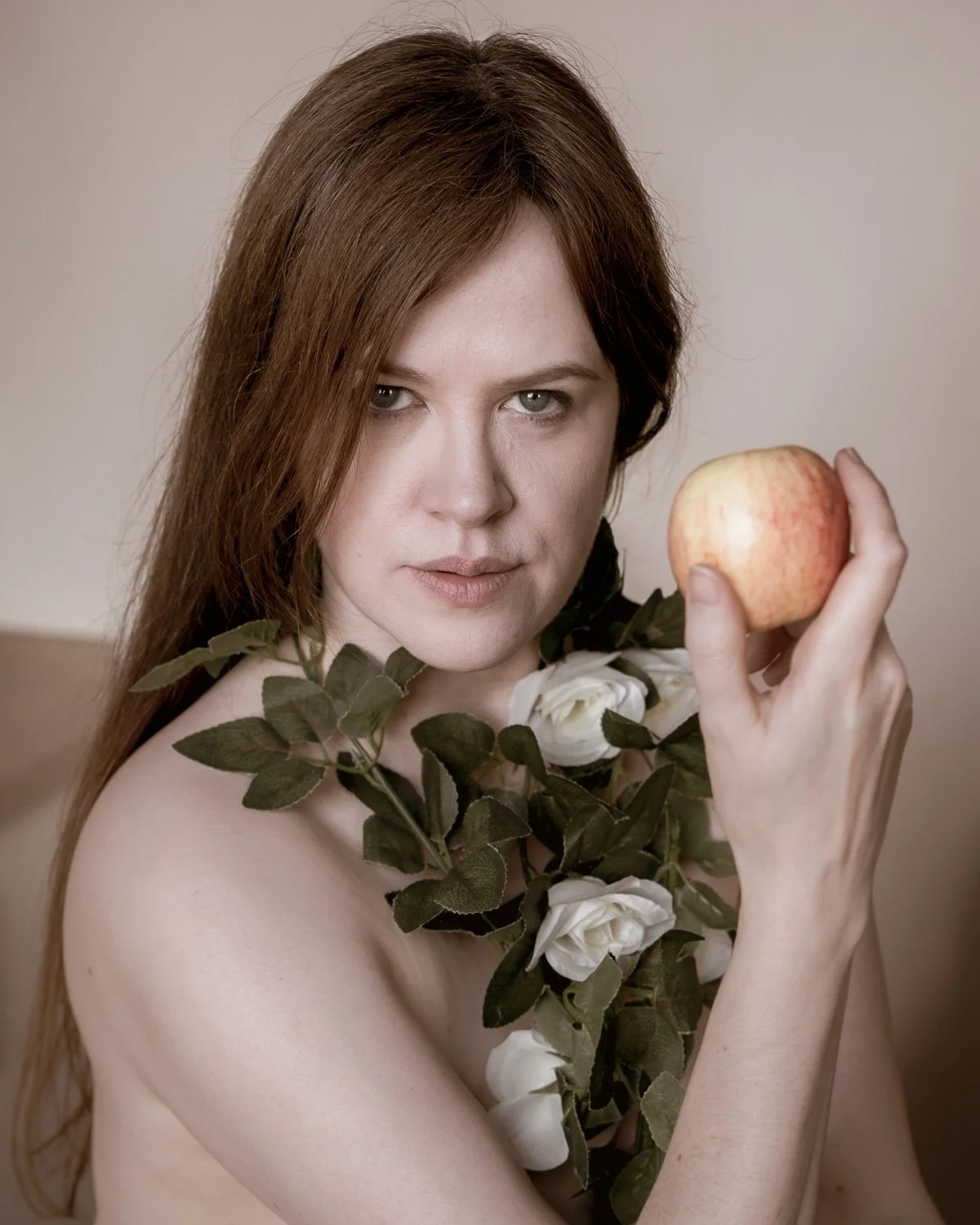 A woman with long auburn hair holding a red and yellow apple near her face, with white roses and green leaves draped over her shoulder.