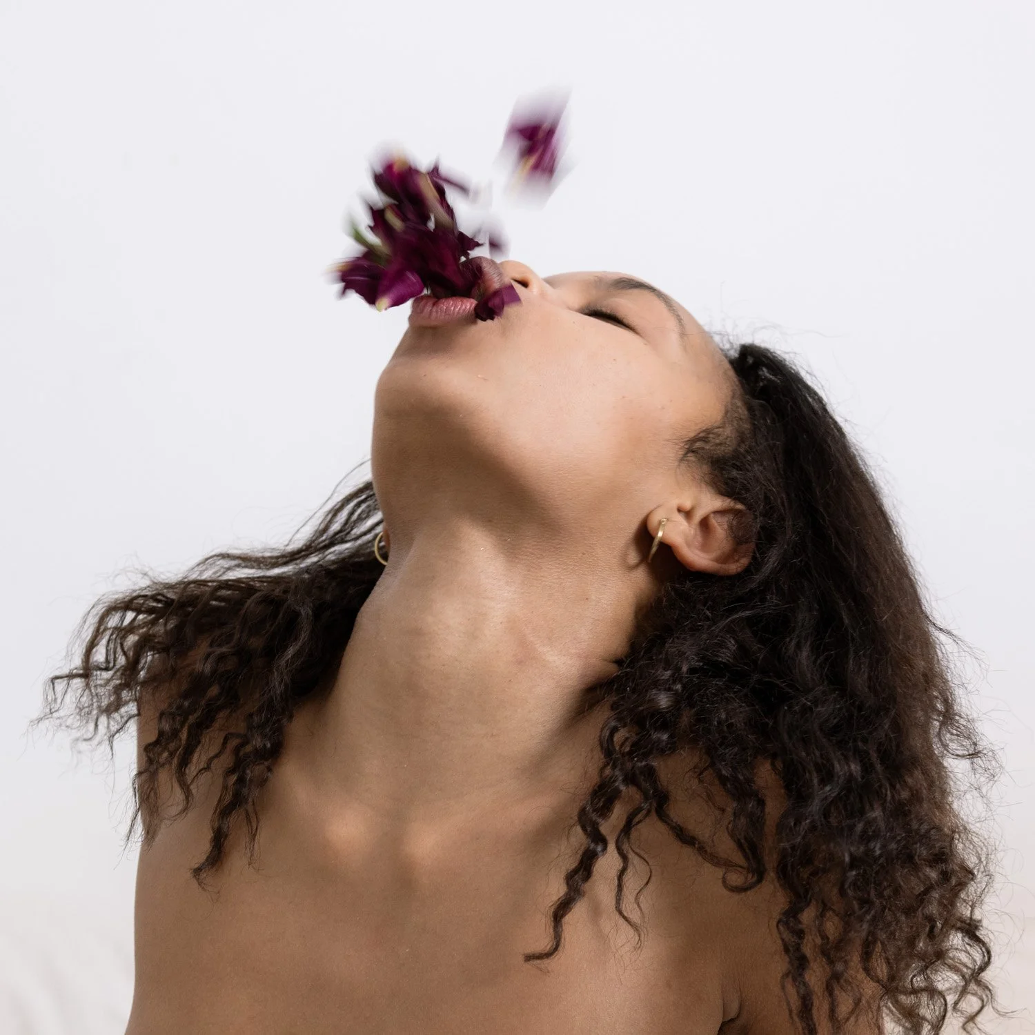 A woman with curly hair and earrings tilting her head back, blowing purple flower petals from her open mouth.