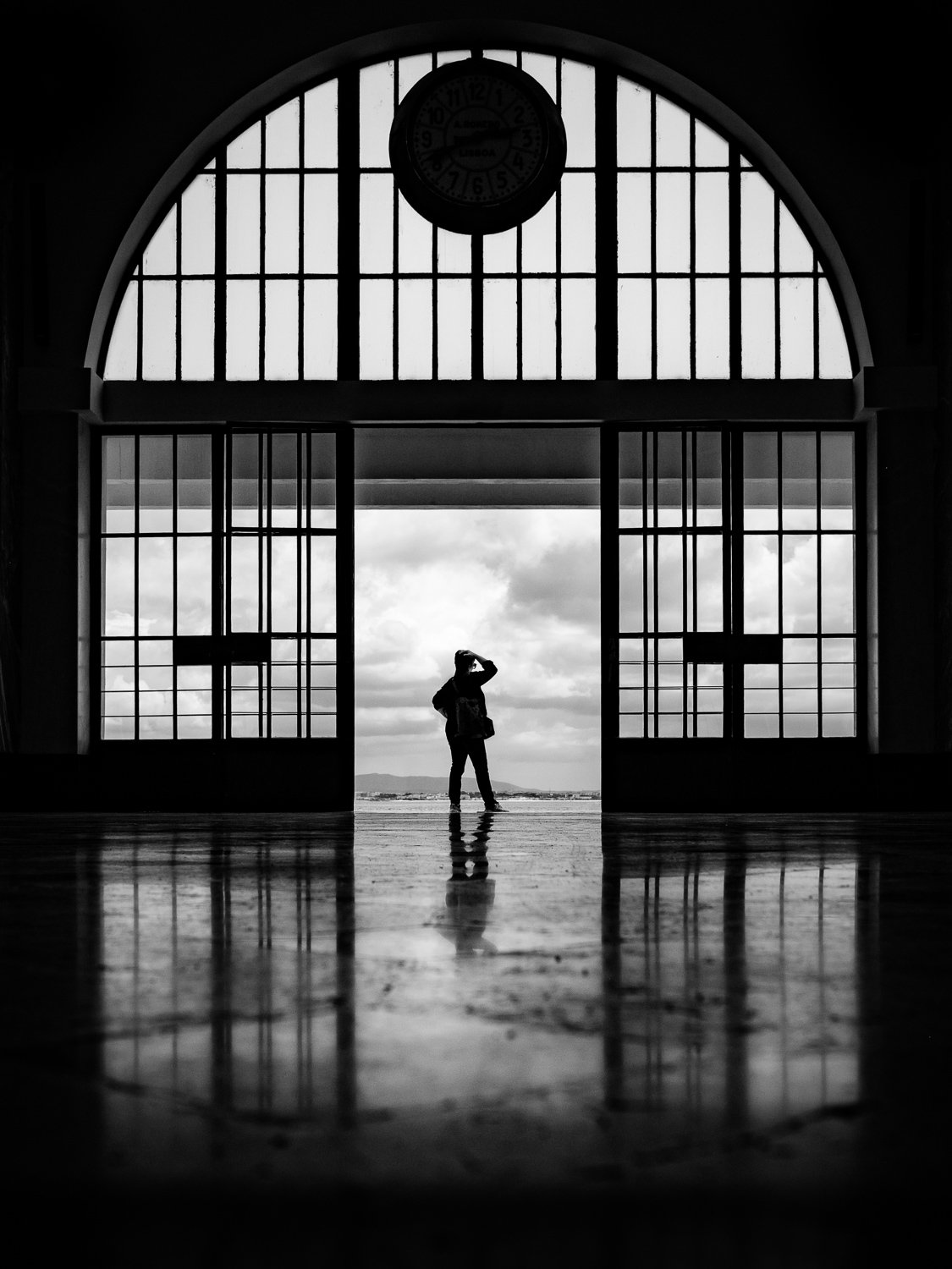 Black and white photo of a person standing near large open doors with a view of the sky and landscape outside, inside a building with a large arched window and a clock at the top.
