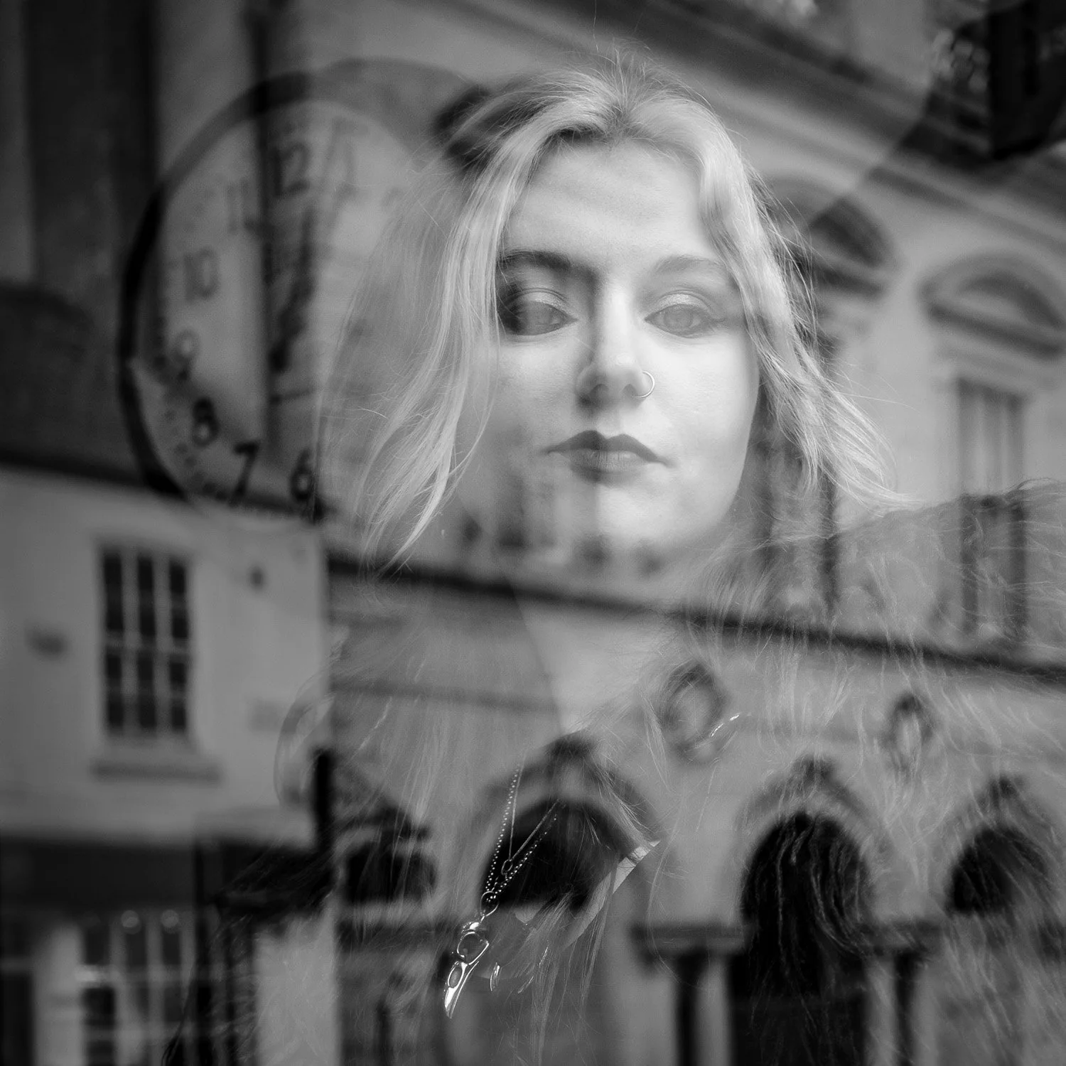 A woman with blonde hair, nose ring, and dark makeup, looking through a window with reflections of buildings and a clock.