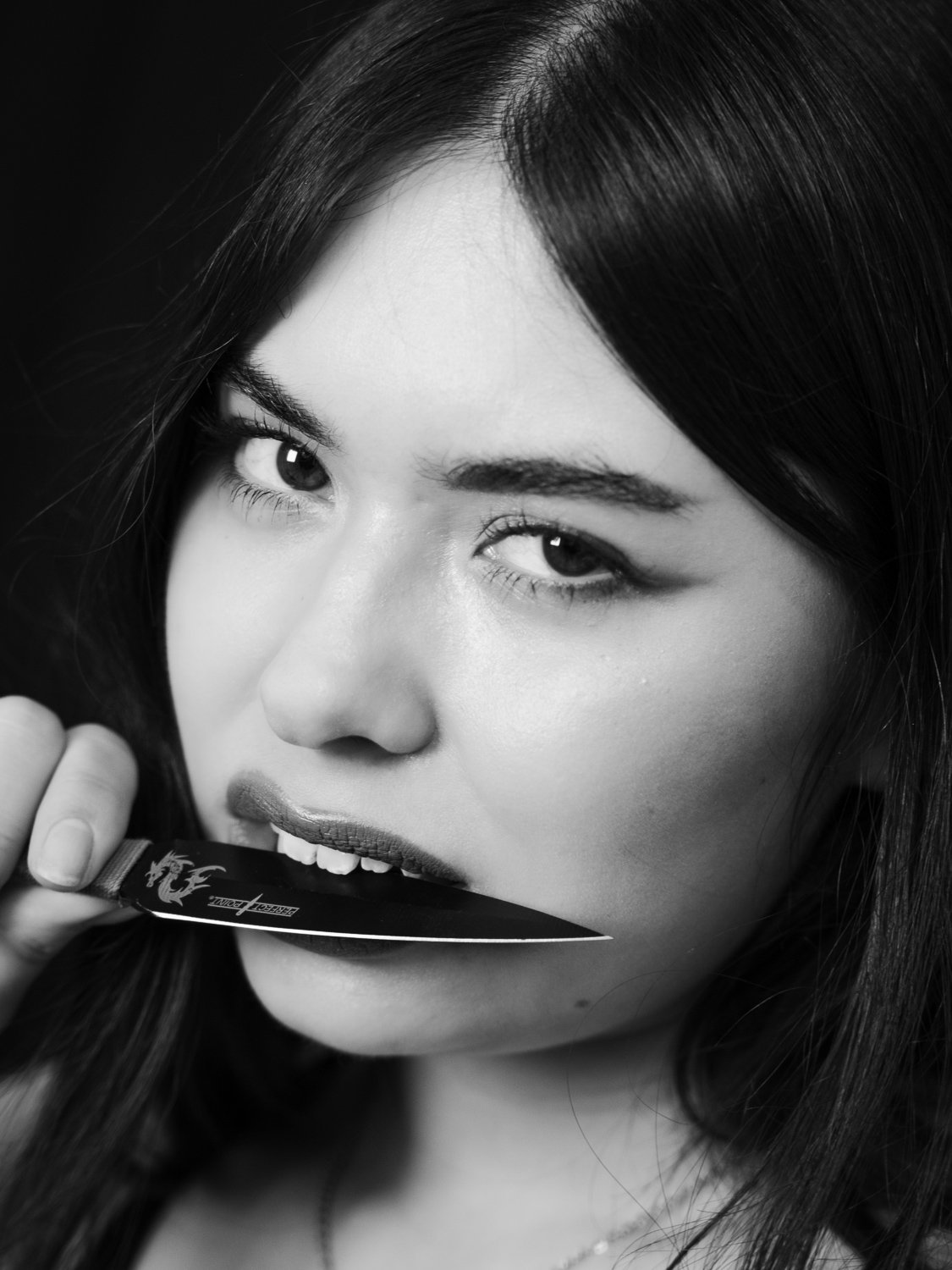 Black and white close-up of a woman with long hair biting a knife with a dragon design.