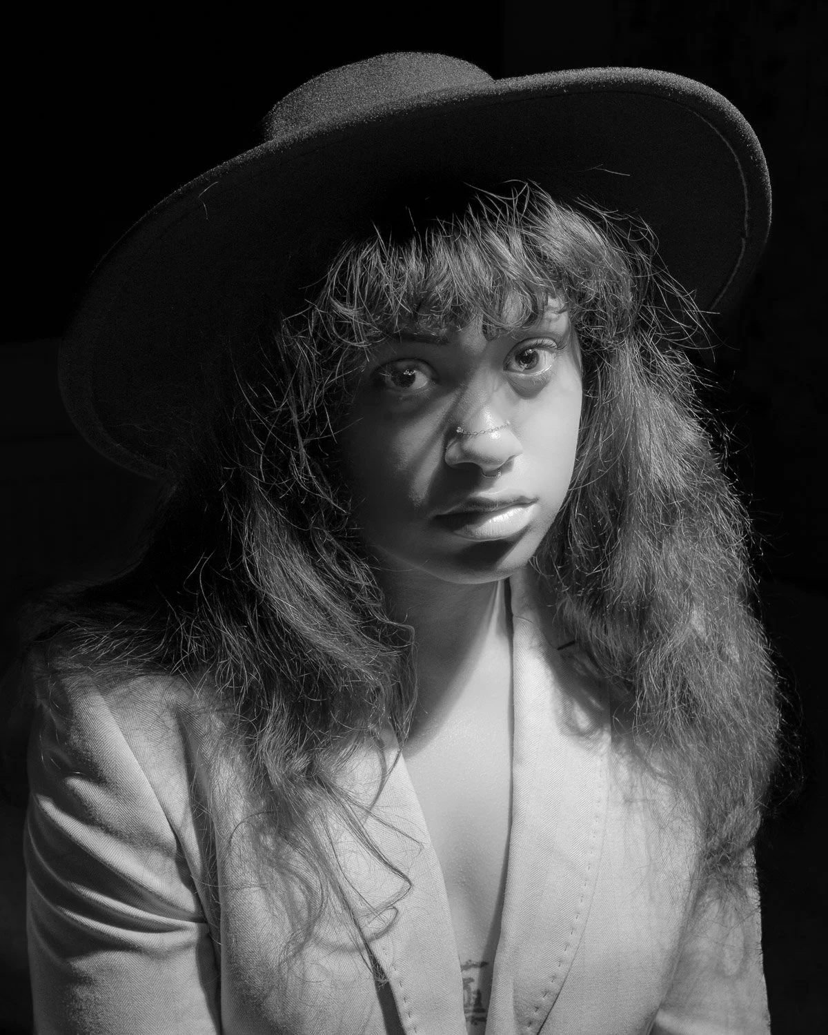 Black and white portrait of a woman wearing a wide-brimmed hat, with curly hair, light-colored blazer, and a septum piercing, looking directly at the camera.