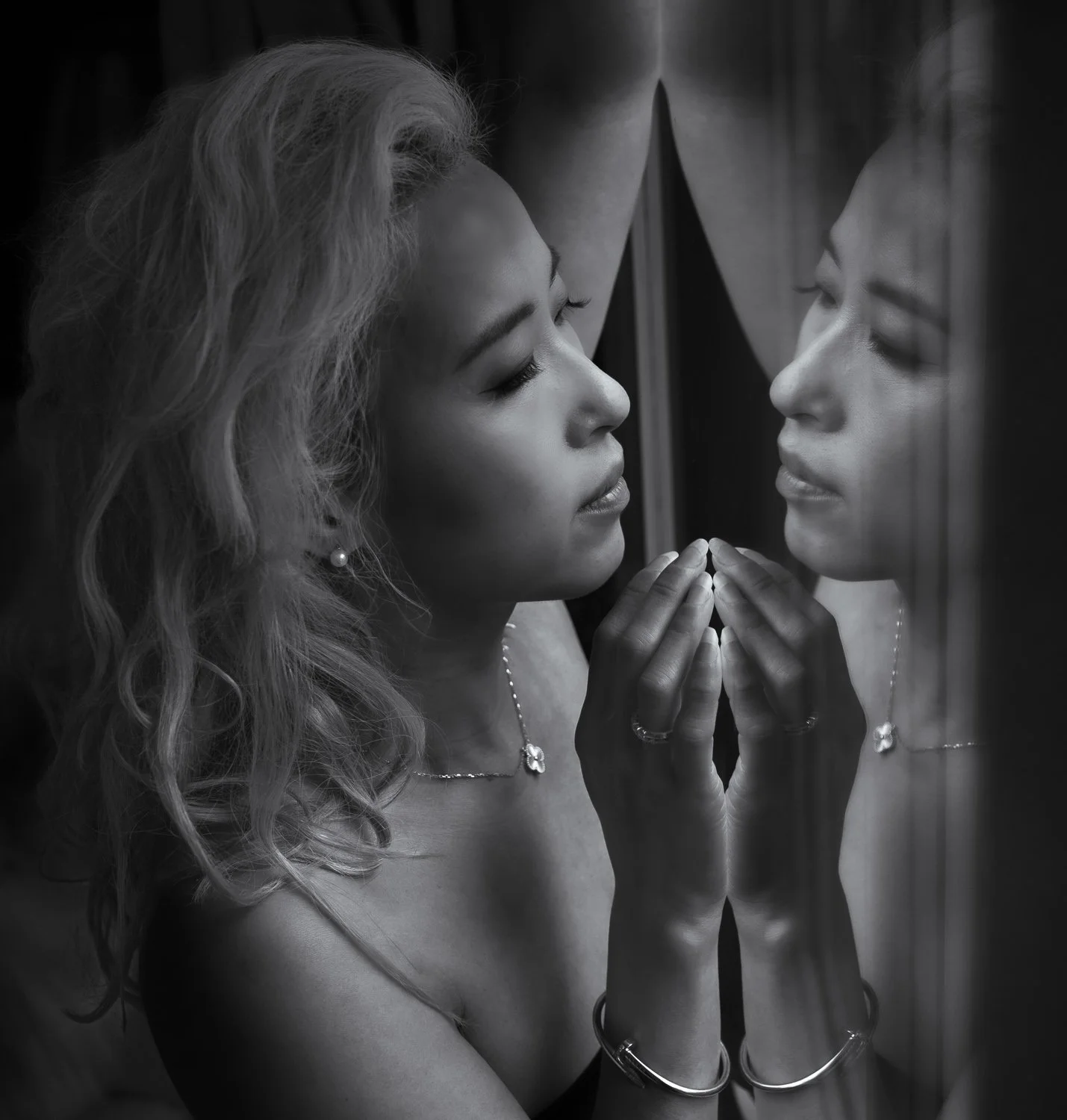 A woman with wavy hair wearing jewelry is looking at her reflection on a glass surface, with her hands gently pressed against the glass.