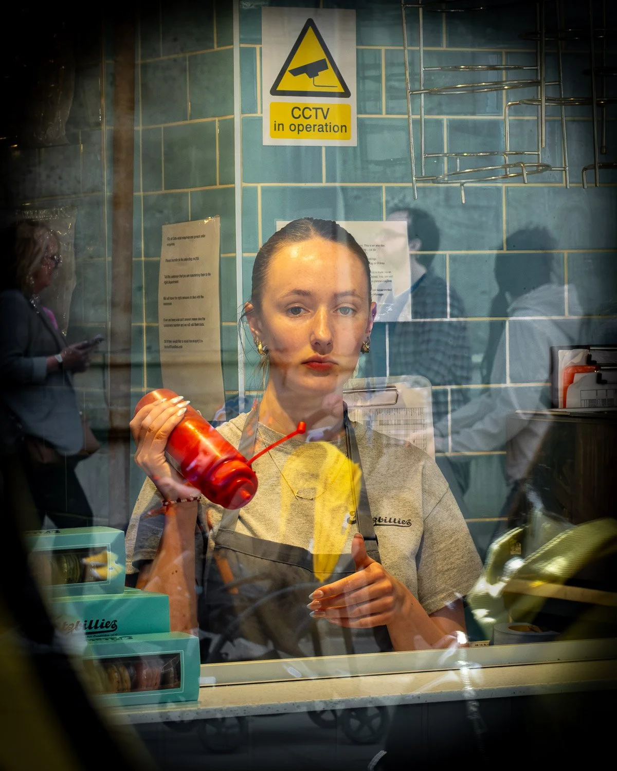 A woman in a cafe or food counter behind a glass window, holding a red ketchup bottle and warning yellow cloth, with a serious facial expression. The background shows a dark blue tiled wall with a CCTV sign, and reflections of other people and object