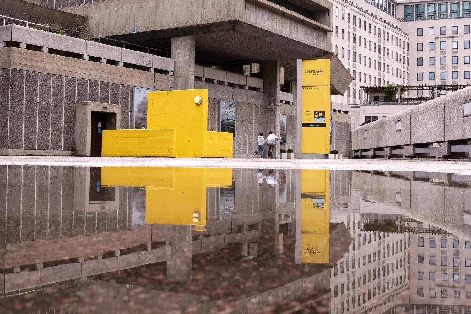 Modern urban building facade with gray concrete and glass, yellow signage and structures, and a reflective water puddle in the foreground reflecting the scene.