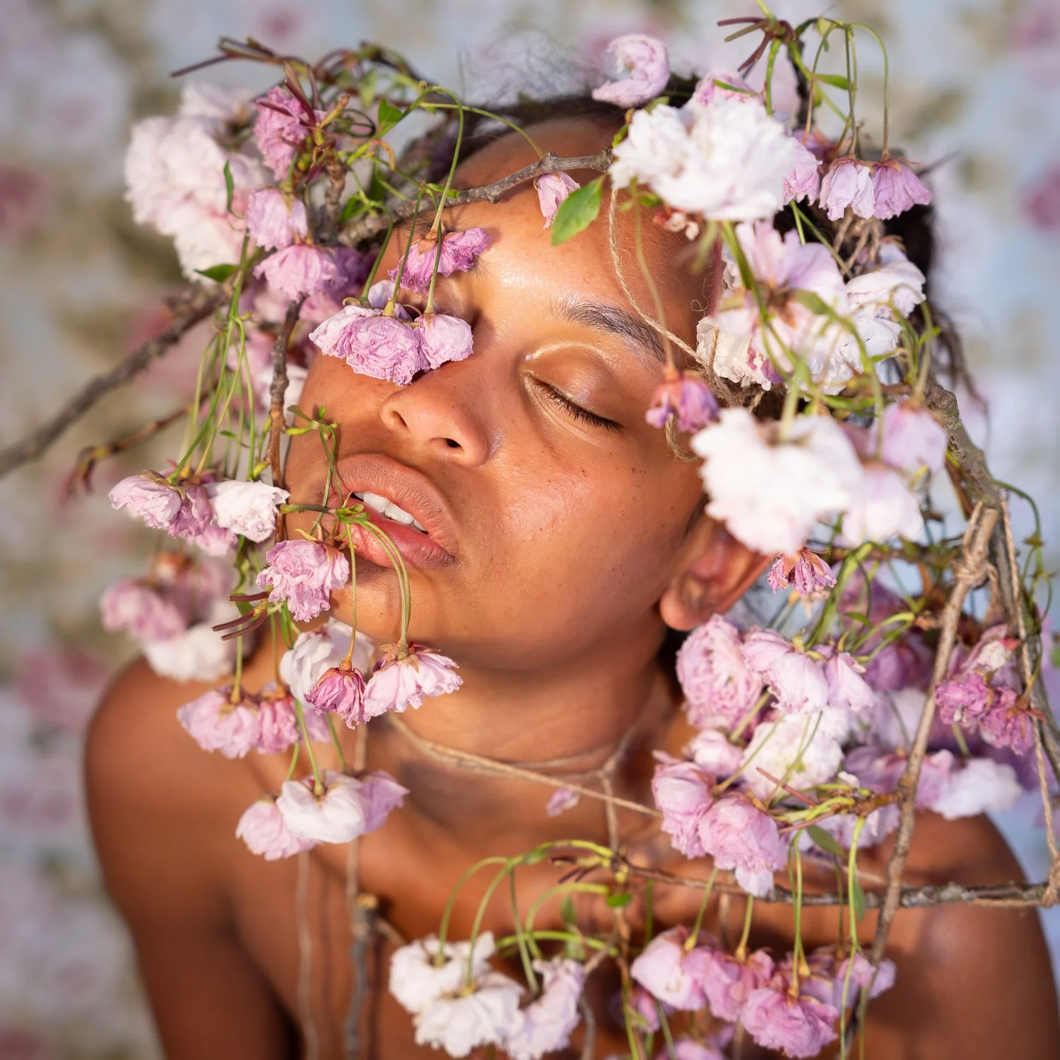 Close-up of a woman with closed eyes, surrounded by pink and white flowers and branches that create a crown around her head, conveying serenity and connection with nature.