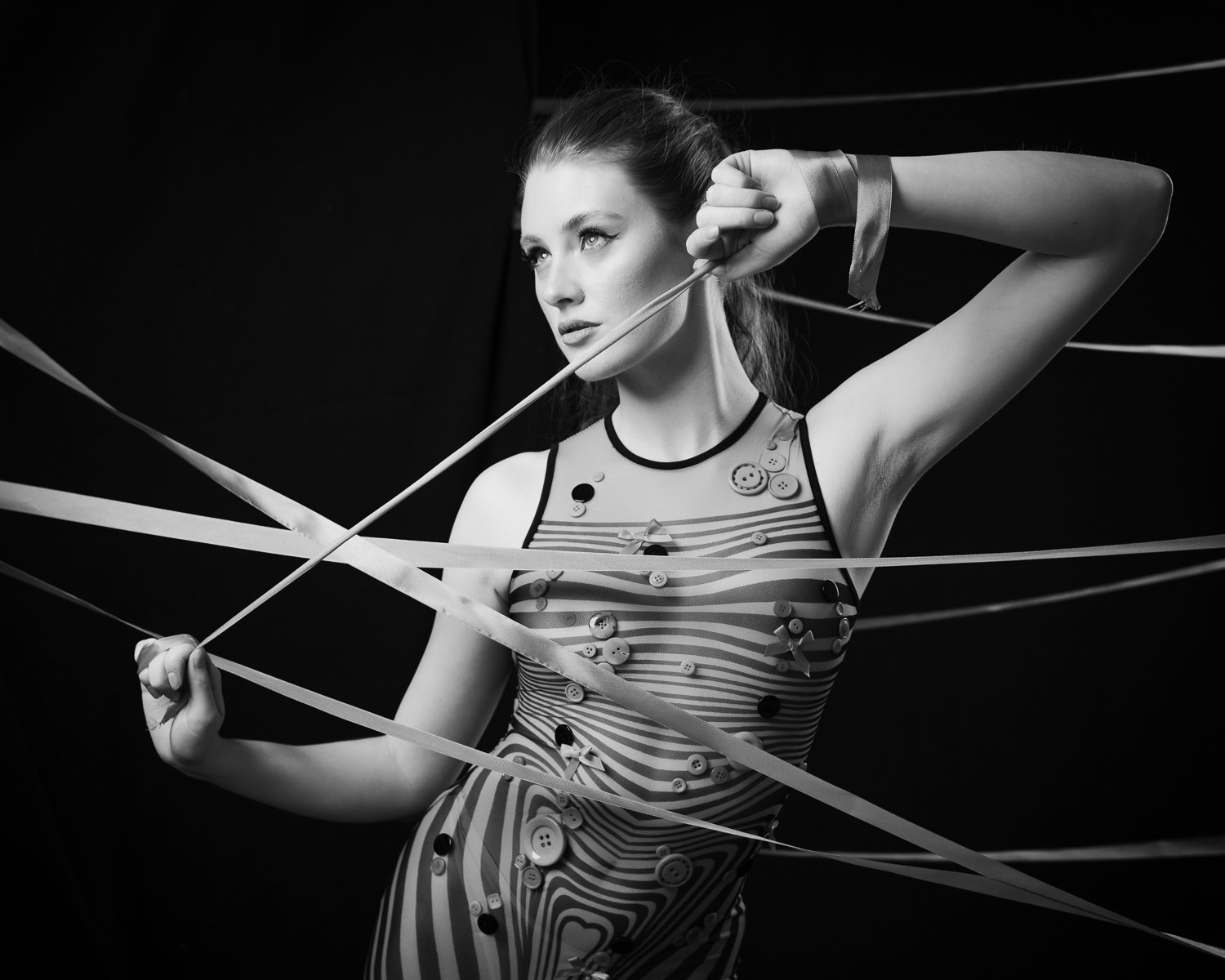 Black and white photo of a woman with buttons and ribbons on her striped dress, holding ribbons and a stick, with a dark background.