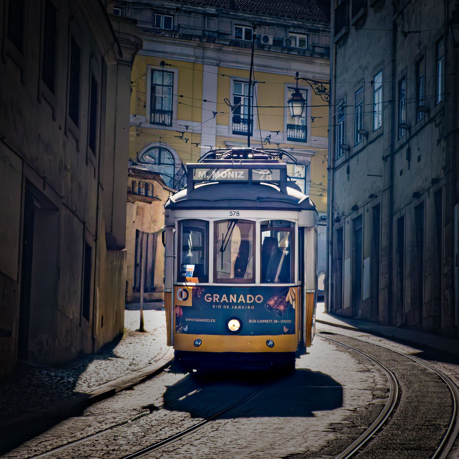 A yellow tram traveling on a narrow street, surrounded by old buildings in a European city at night.