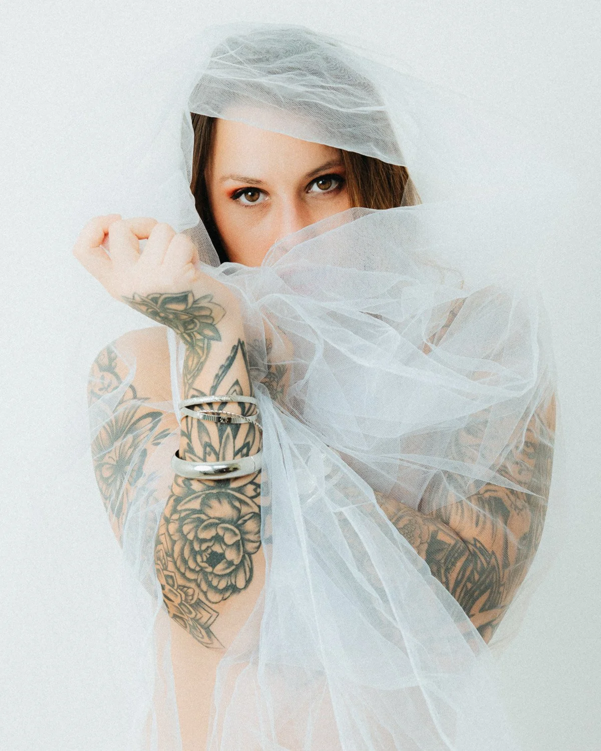 A woman with tattoos on her arms and shoulders, partially covered with sheer white fabric, looking directly at the camera with her face partially obscured by the fabric.