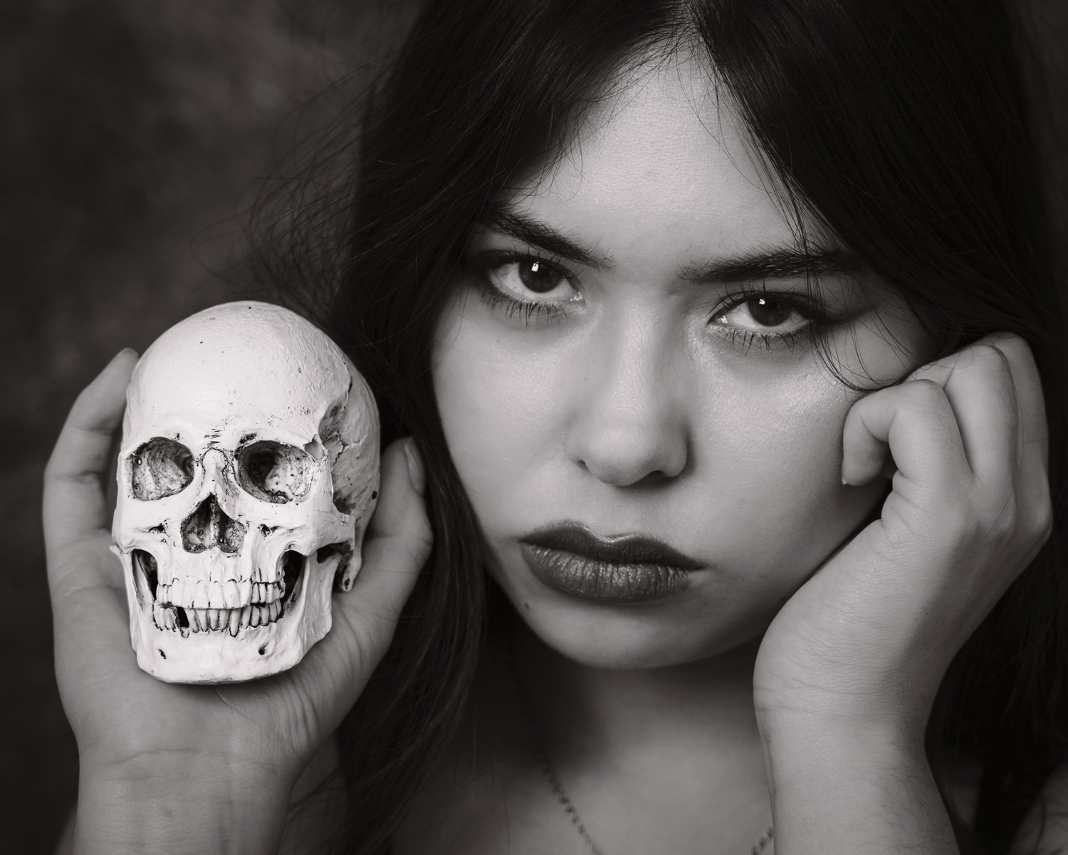 A black and white photo of a woman with dark hair holding a human skull close to her face, looking directly at the camera with intense eyes.