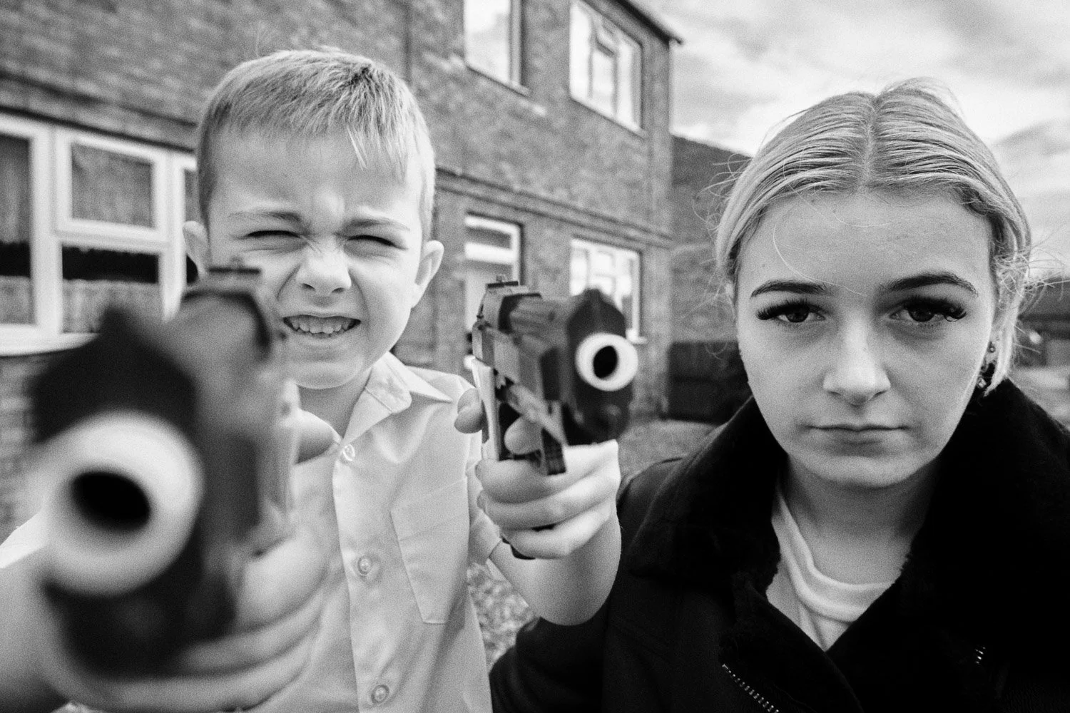 A young boy and a young woman pointing toy guns at the camera, outside a brick building, with serious expressions on their faces. Black and white photograph.