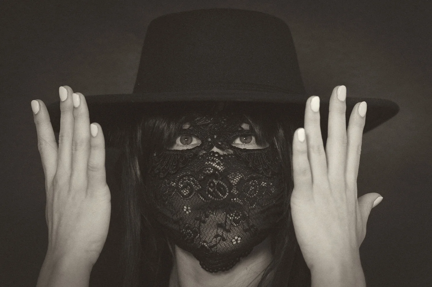 A woman wearing a black wide-brimmed hat and a black lace face mask, looking directly at the camera.