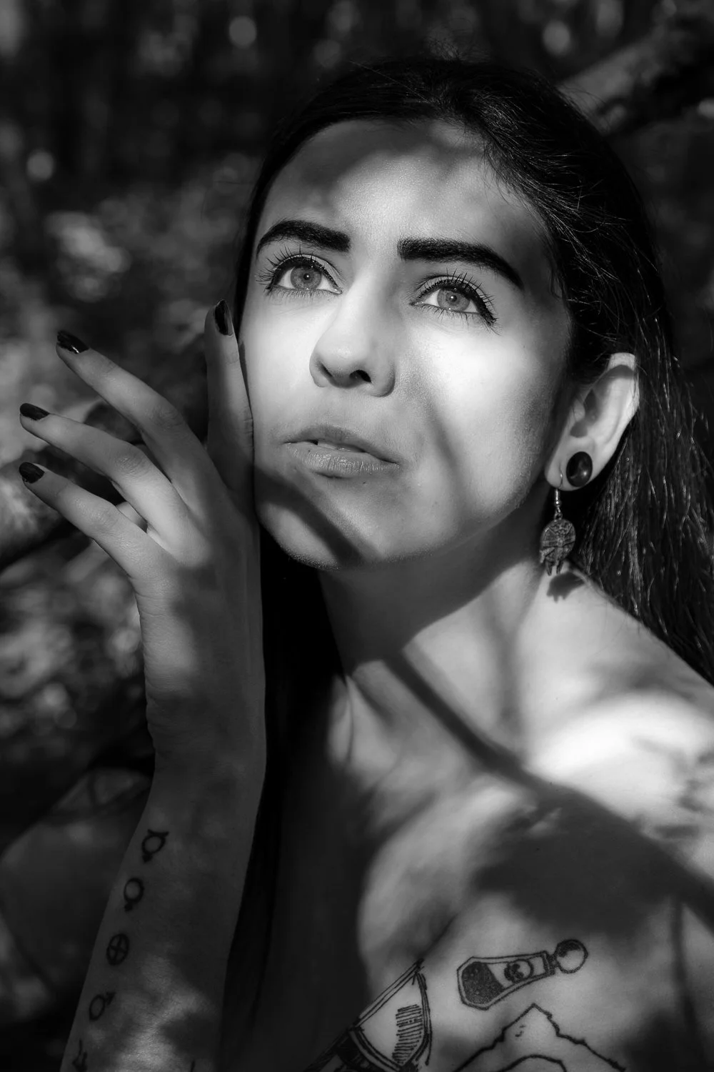 Close-up black and white portrait of a young woman with dark hair, wearing earrings, and tattoos on her arm and shoulder, outdoors with blurred natural background, looking thoughtfully upwards.