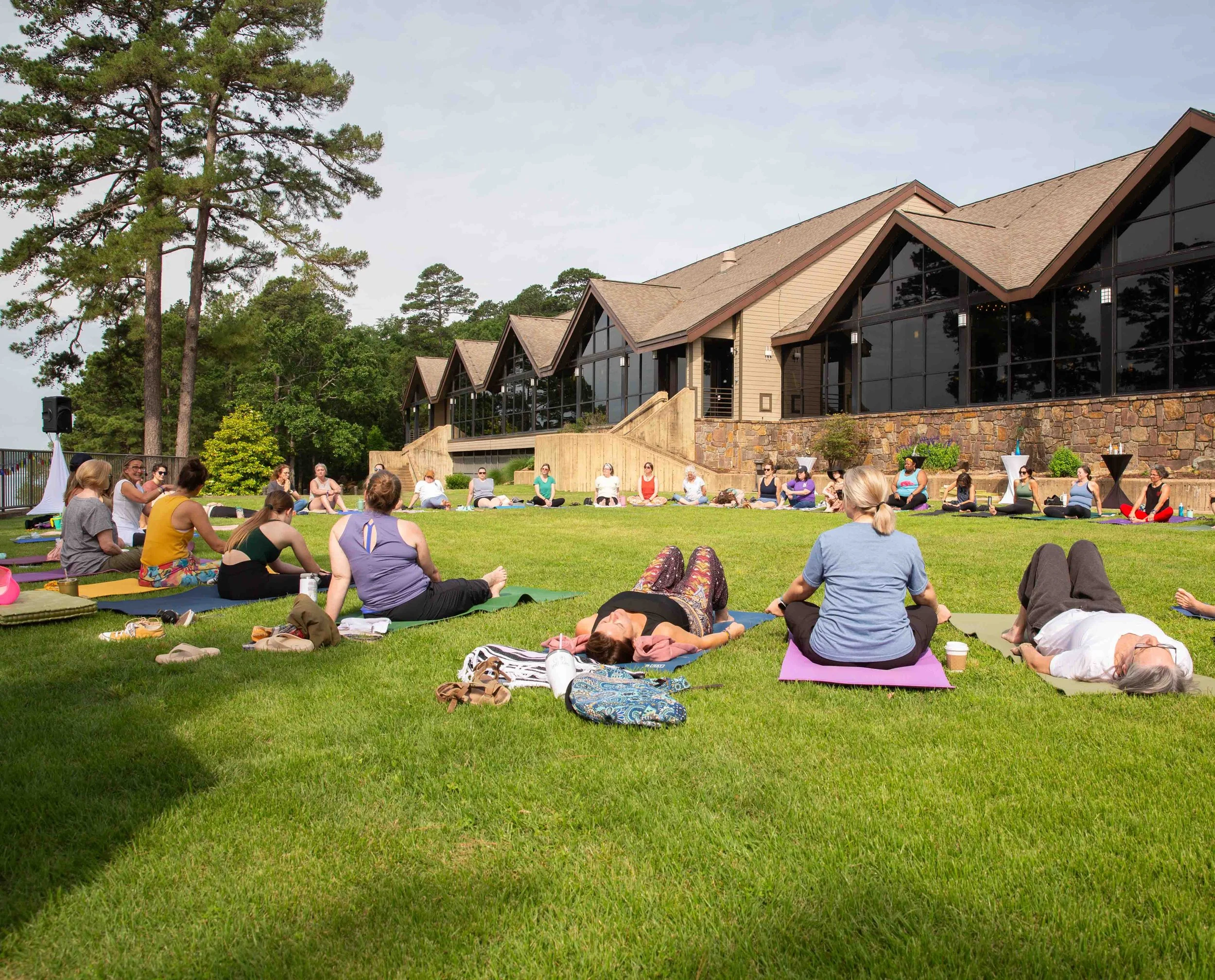A group of people practicing yoga on a grassy lawn in front of a modern building with large glass windows and a stone foundation, surrounded by trees.