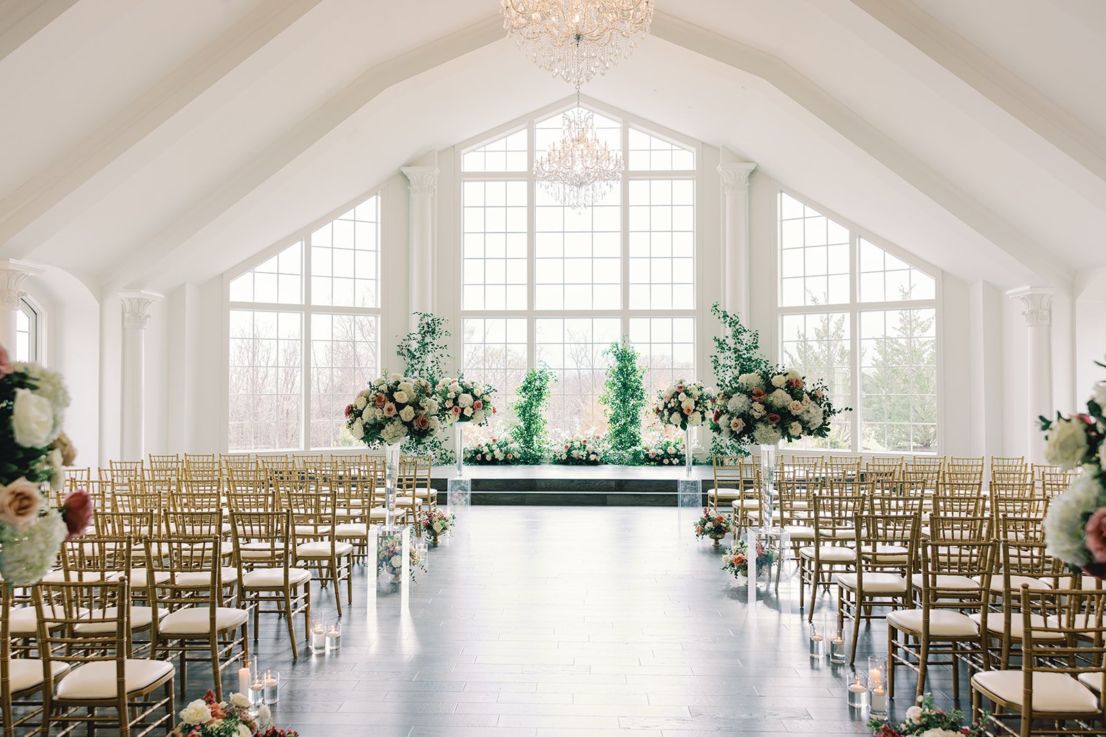 Beautifully decorated indoor wedding venue with large windows, floral arrangements, and rows of chairs facing an altar with candles.