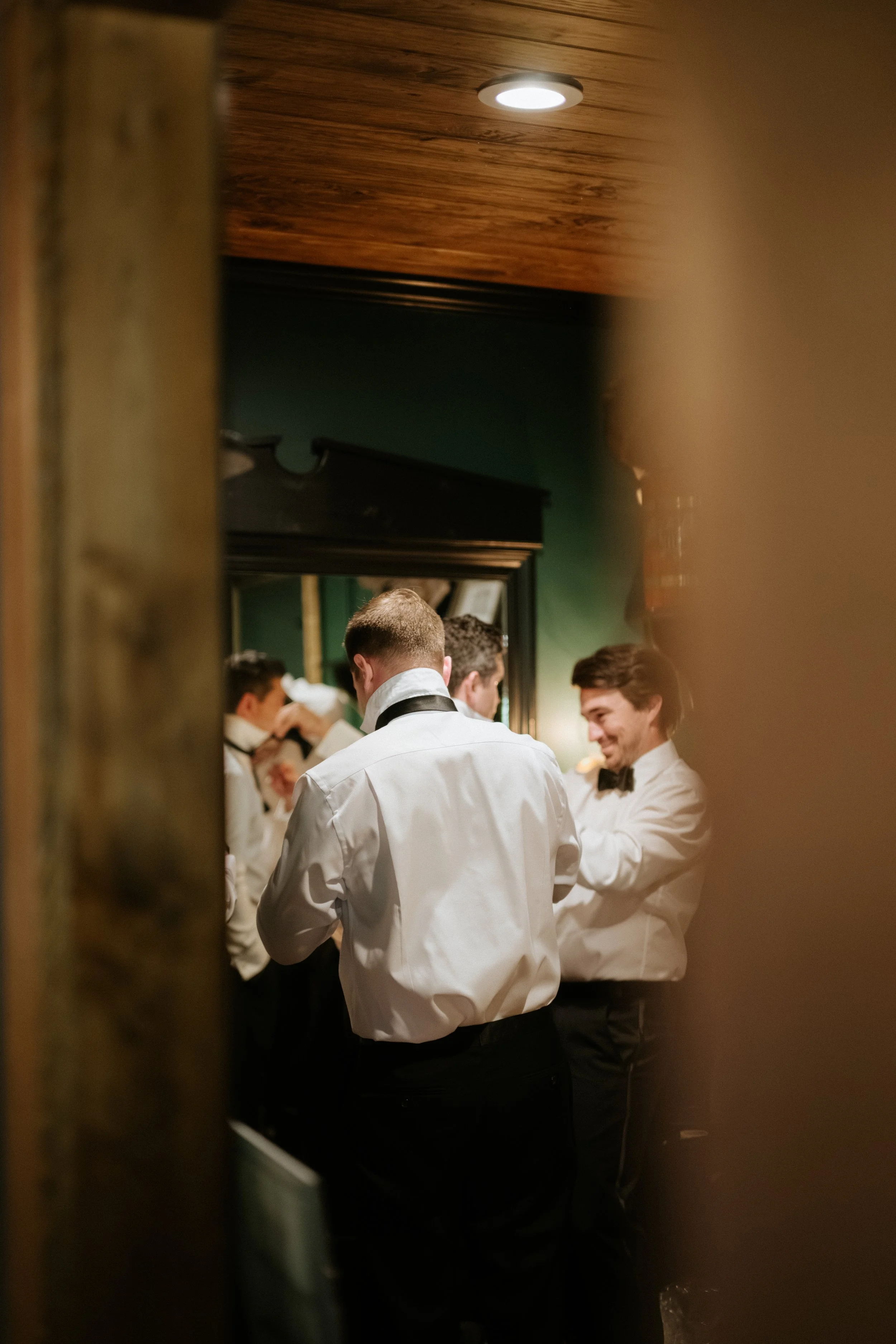 Groom and groomsmen adjusting their tuxedos in a mirror, seen through a narrow gap in the doorway, in a dimly lit room with green walls and wooden ceiling.