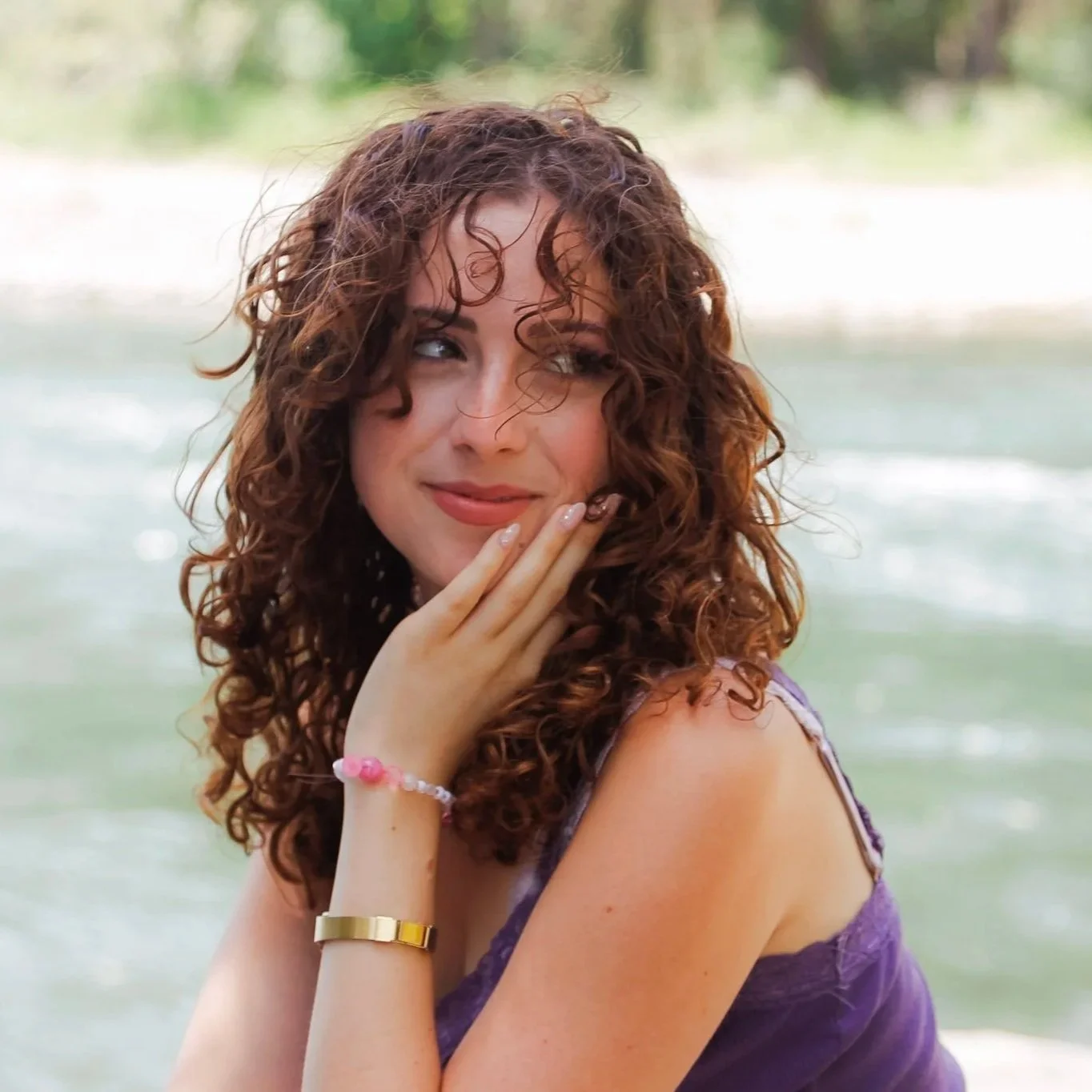 A young woman with curly auburn long hair looking over her left shoulder towards the right of the photo. Her left hand is resting on her chin. She is wearing two bracelets and a purple tank top. Behind her is a blurry river and riverbank.