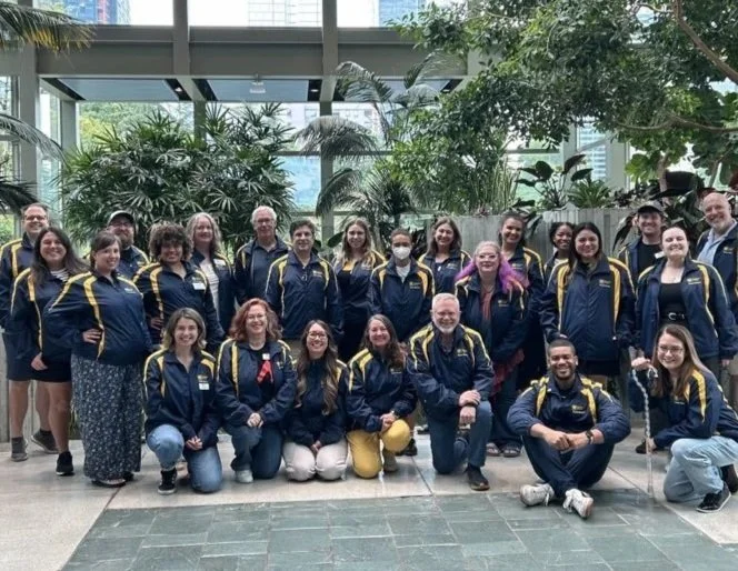 Group of 21 people in navy blue jackets with yellow accents posing together in a lush indoor botanical garden with large windows and green plants.