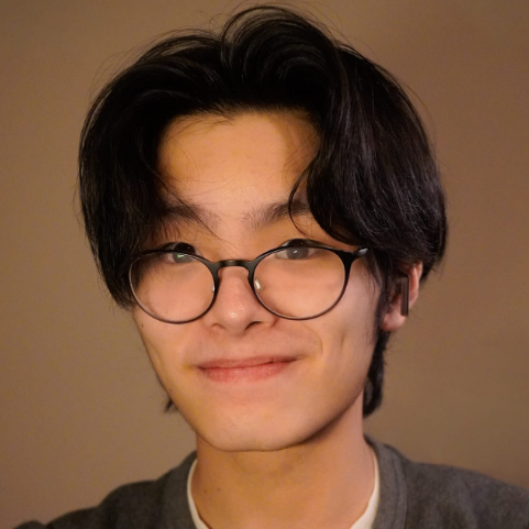 A young man wearing with medium length black hair and brown eyes smiling at the camera. He is wearing black, round lens glasses and a dark grey shirt. Behind him is a brown background.