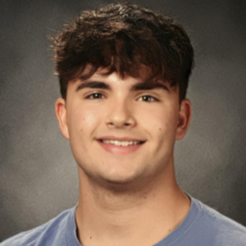 A young man with short brown hair and brown eyes. He is smiling and wearing a blue shirt. Behind him is a dark grey-brown background.