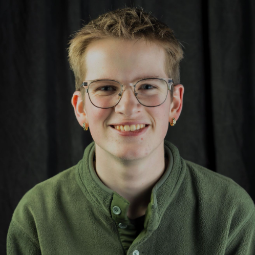 A young man with short light brown hair smiling at the camera. He has earrings in both ears and is wearing a green button up shirt and round-rimmed glasses. Behind him is a black background.