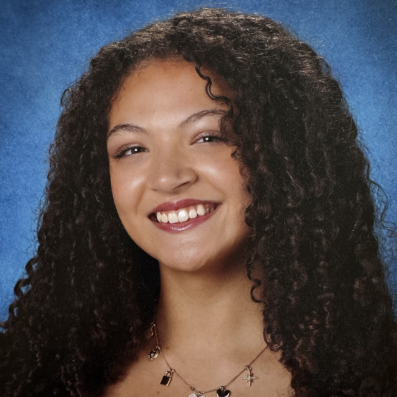 A young woman with long, dark, curly hair and brown eyes. She is smiling and wearing a necklace. Behind her is a blue background.
