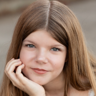 A young woman with straight light brown hair and blue eyes resting her chin on her right hand looking at the camera. Behind her is a medium grey background.