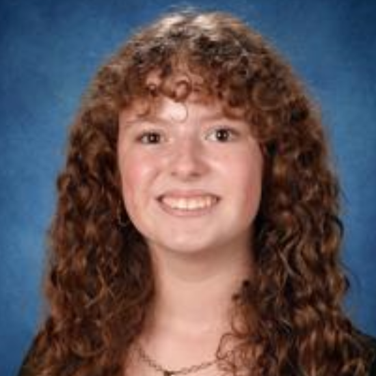 A young woman with long curly brown hair and brown eyes. She is smiling. She is wearing a black top and a necklace. Behind her is a blue background.