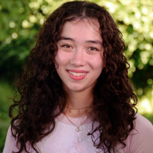 A young woman smiling with dark wavy hair wearing a pink top. A blurred green background.