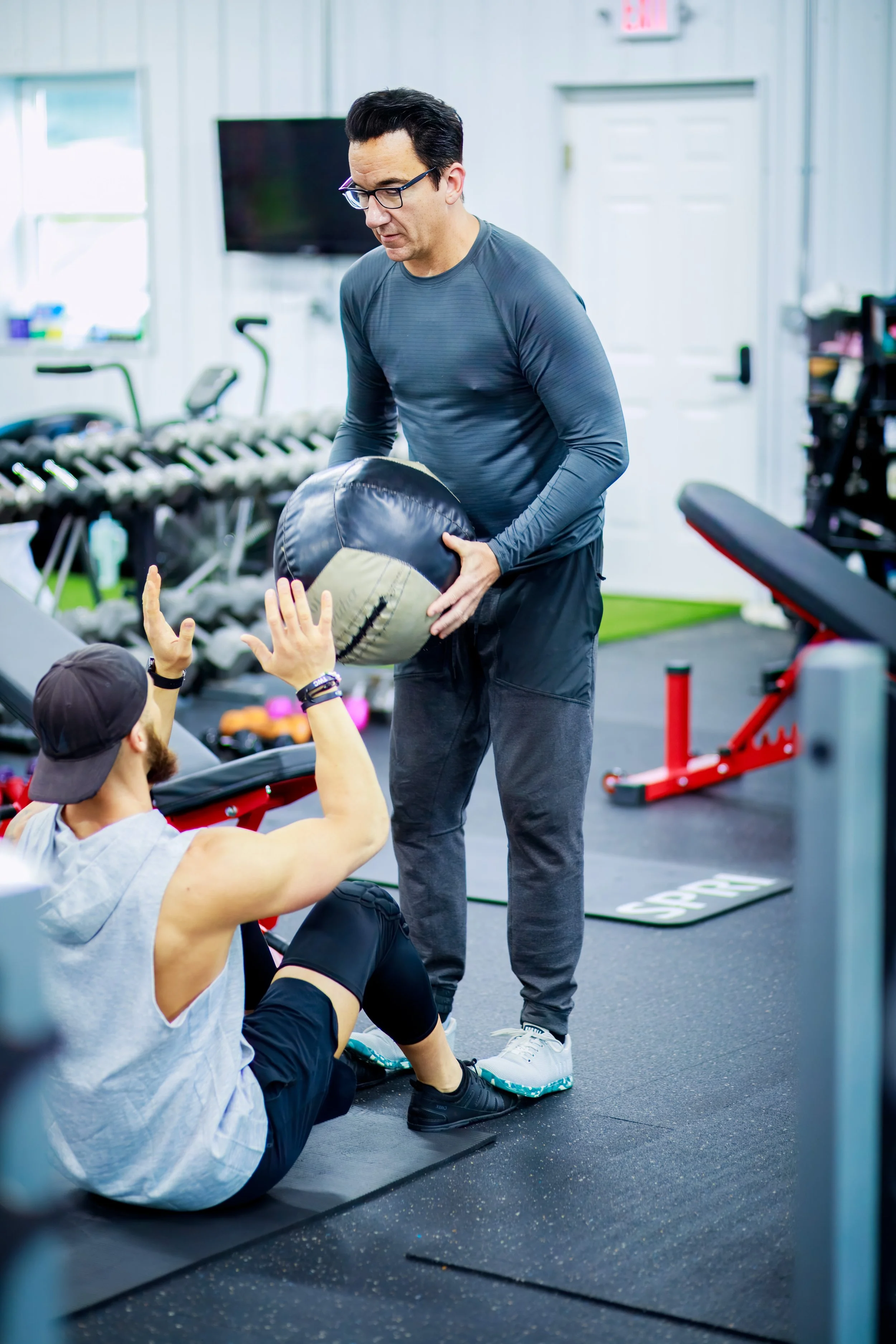 A man handing a medicine ball to a seated man in a gym.