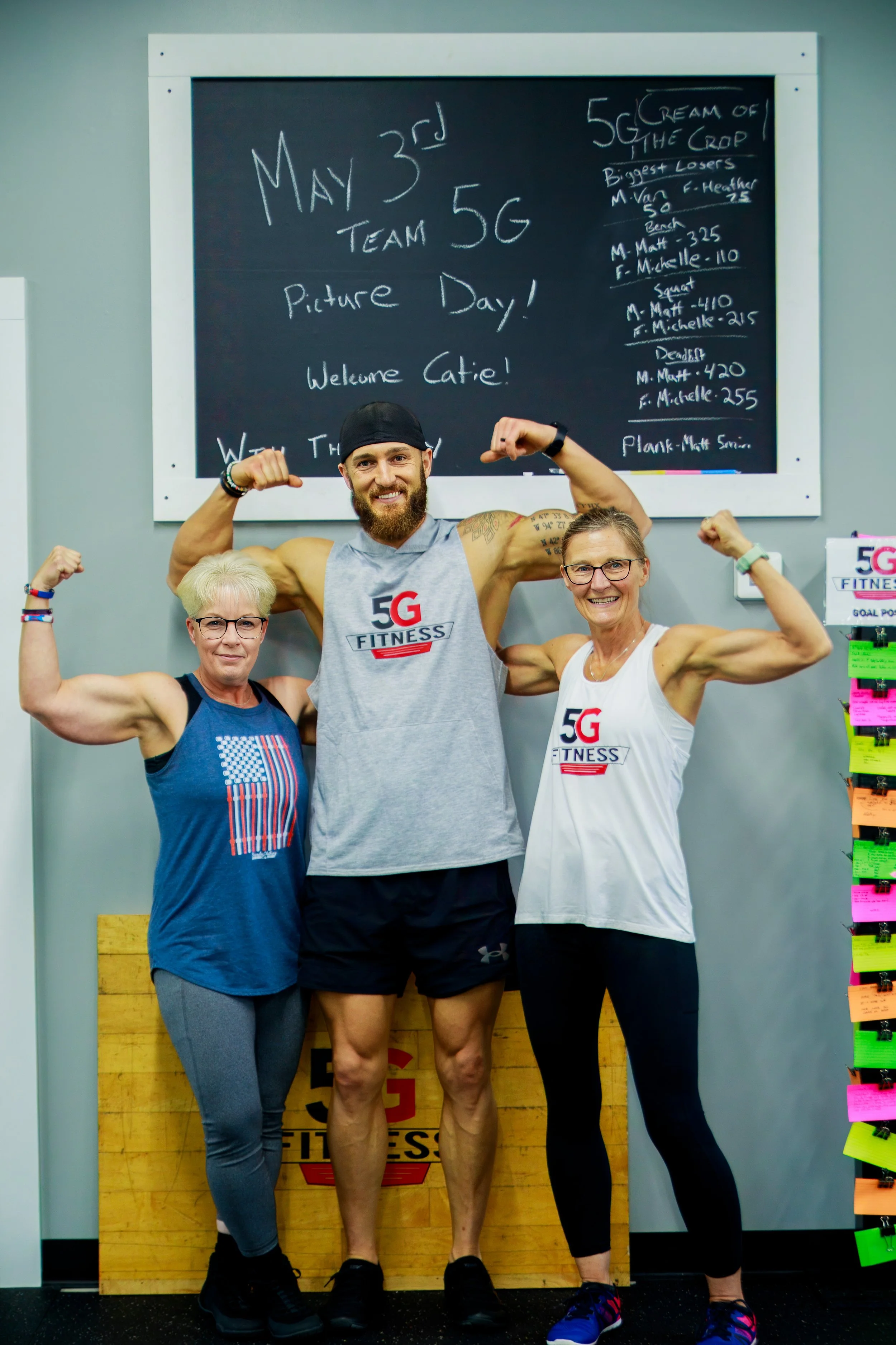 Three people, two women and one man, flexing their arm muscles and smiling at the camera inside a fitness gym. They are wearing fitness attire with the logo '5G Fitness'. The background features a blackboard with workout notes and a colorful chart on the right side.