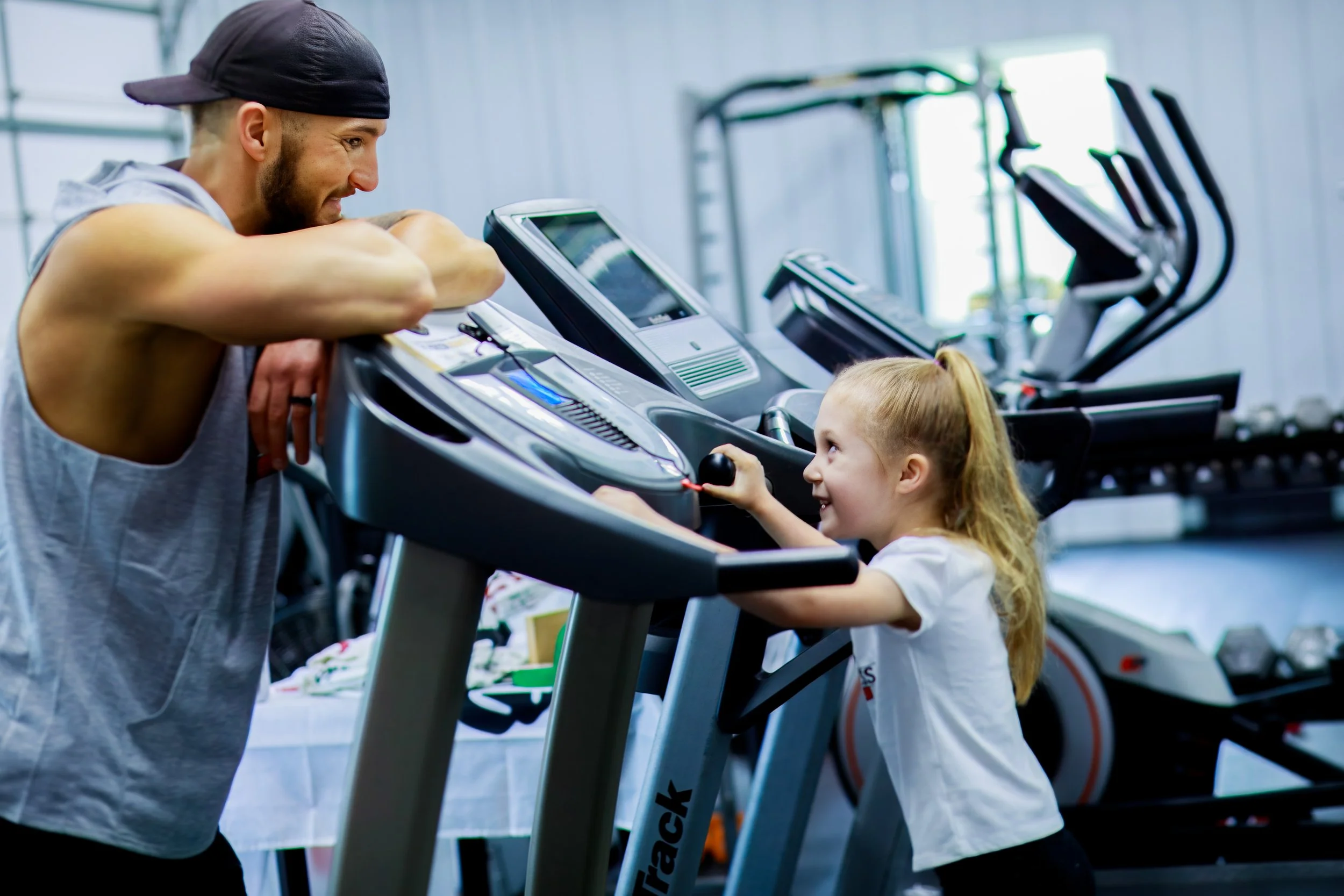 A man and a young girl engaging on a treadmill at a gym. The man is wearing a sleeveless hoodie and a black cap, leaning on the treadmill, while the girl, with a ponytail and wearing a white t-shirt, is holding the treadmill's handle and smiling up a