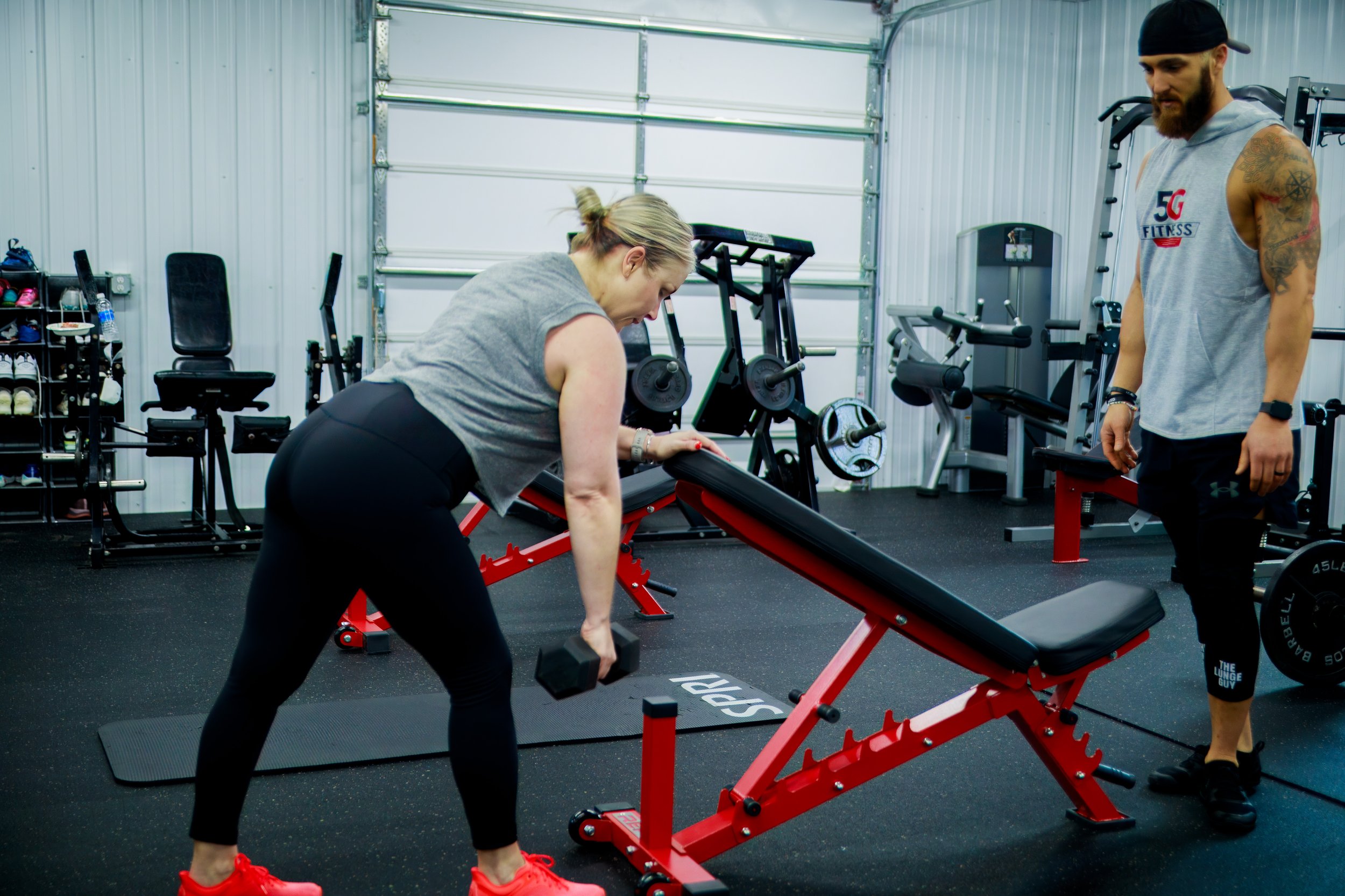 A woman lifting a dumbbell in a gym under the supervision of a male trainer. The woman is in a bent-over position using an adjustable workout bench, with gym equipment and a storage rack in the background.