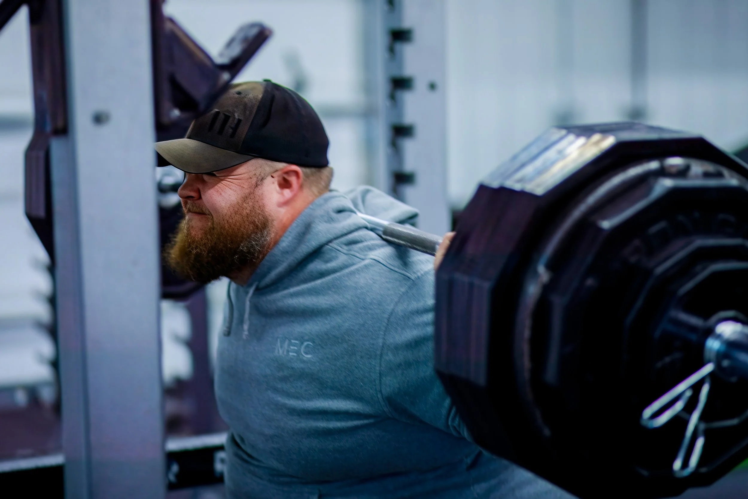 Man with beard wearing a black and beige cap and grey hoodie, lifting a barbell with heavy weights in a gym.