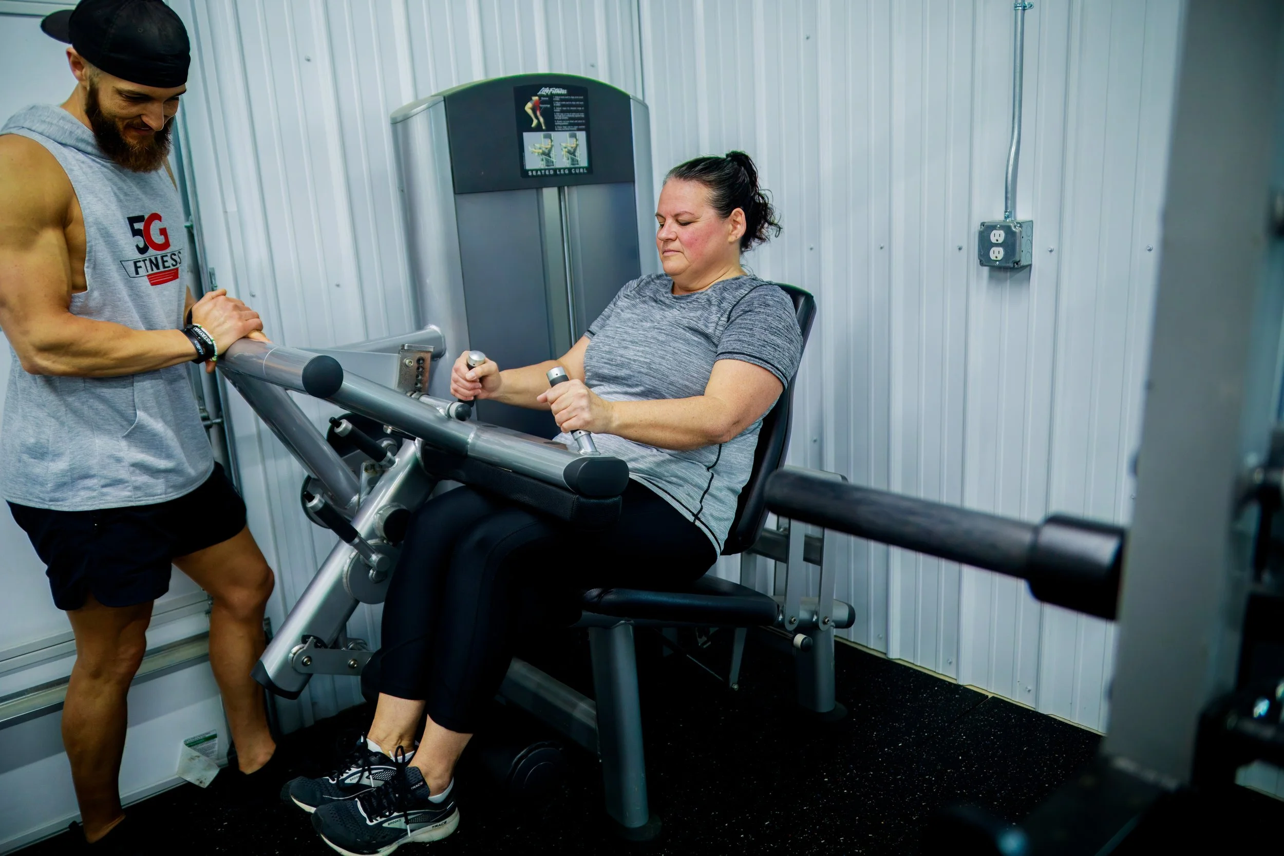 A woman in athletic clothing using a seated leg curl machine at the gym, assisted by a trainer.