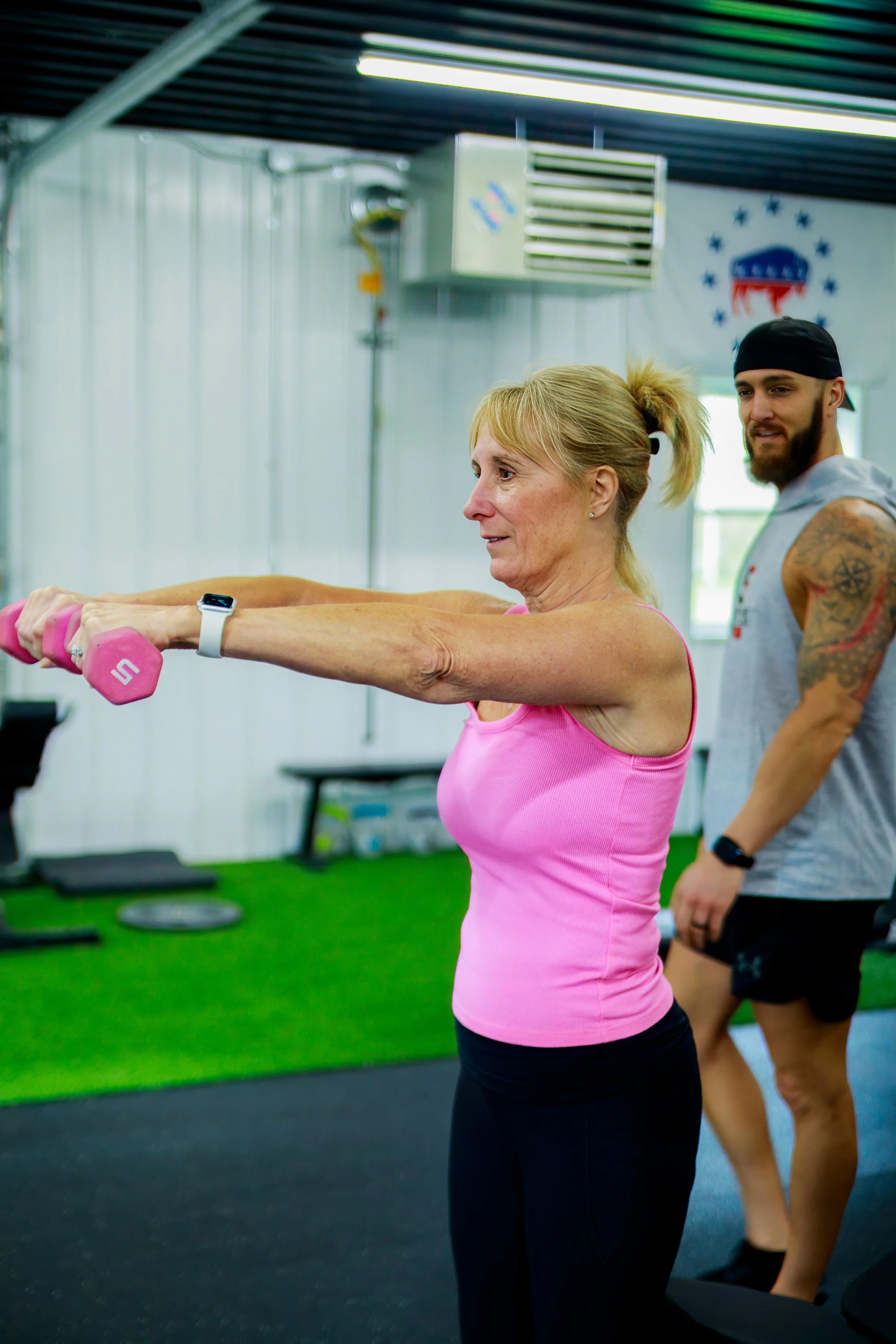 An older woman performs an exercise with pink dumbbells in a gym, supervised by a trainer. The gym has a green artificial turf floor and fitness equipment in the background.
