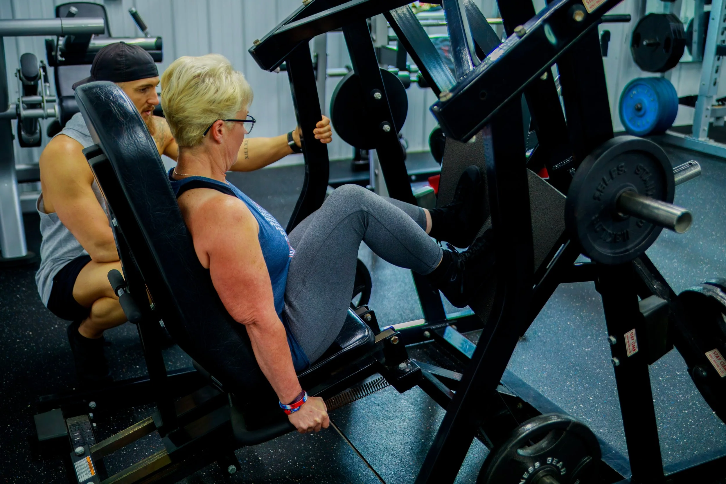 A woman performing a leg press exercise with weights in a gym, supervised by a man.