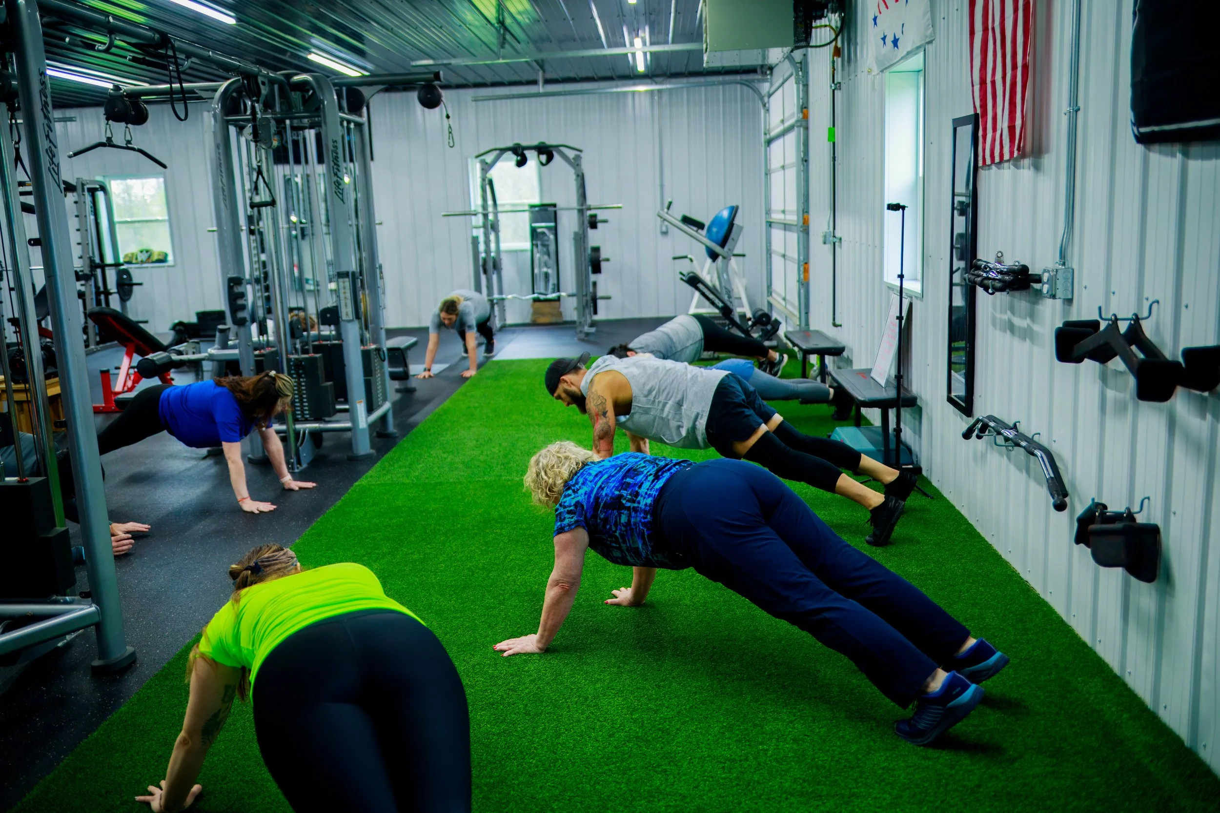 People doing push-ups on artificial turf in a gym with workout equipment and American flag on the wall.