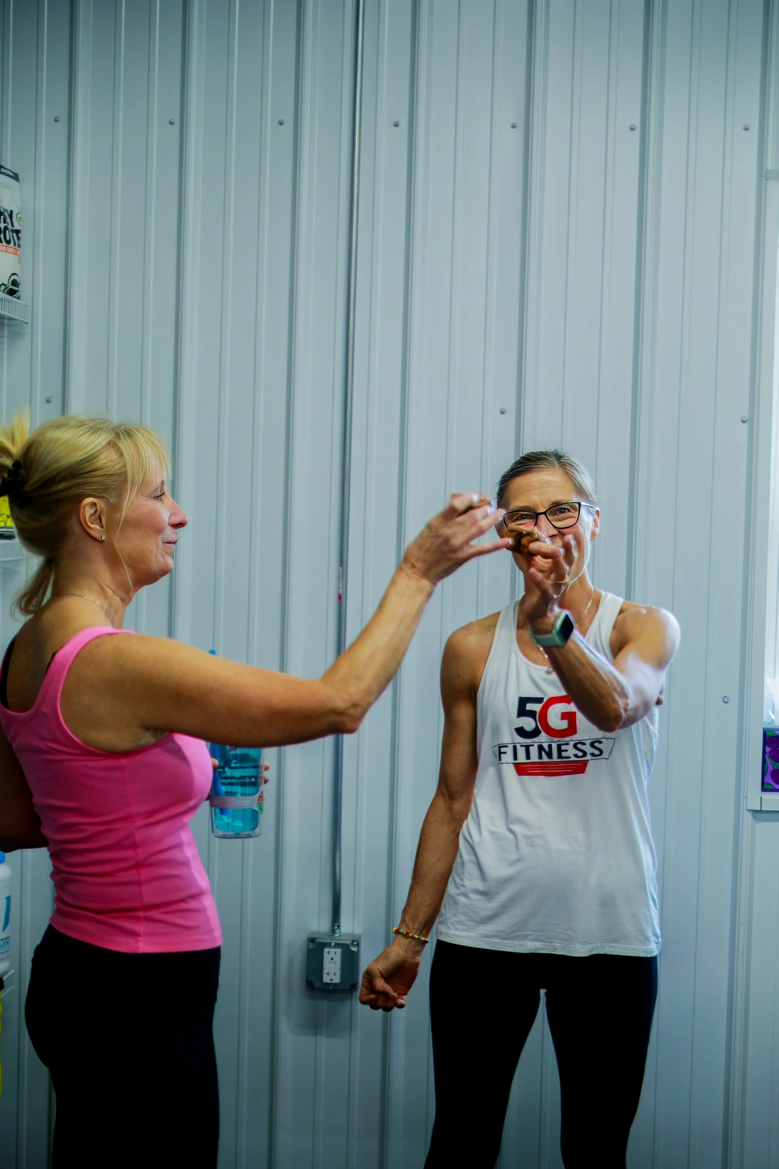 Two women in a gym exchanging a cookie, one is an older woman in a pink tank top and the other a middle-aged woman in a white tank top with 5G FITNESS written on it.