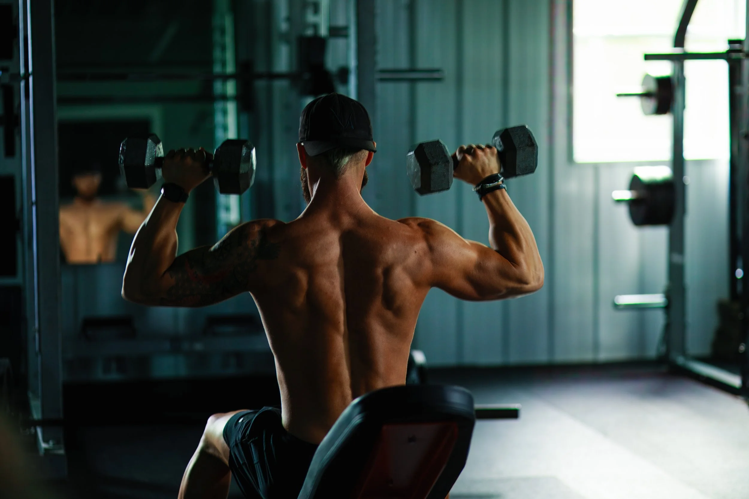 A muscular man lifting dumbbells in a gym, viewed from behind.
